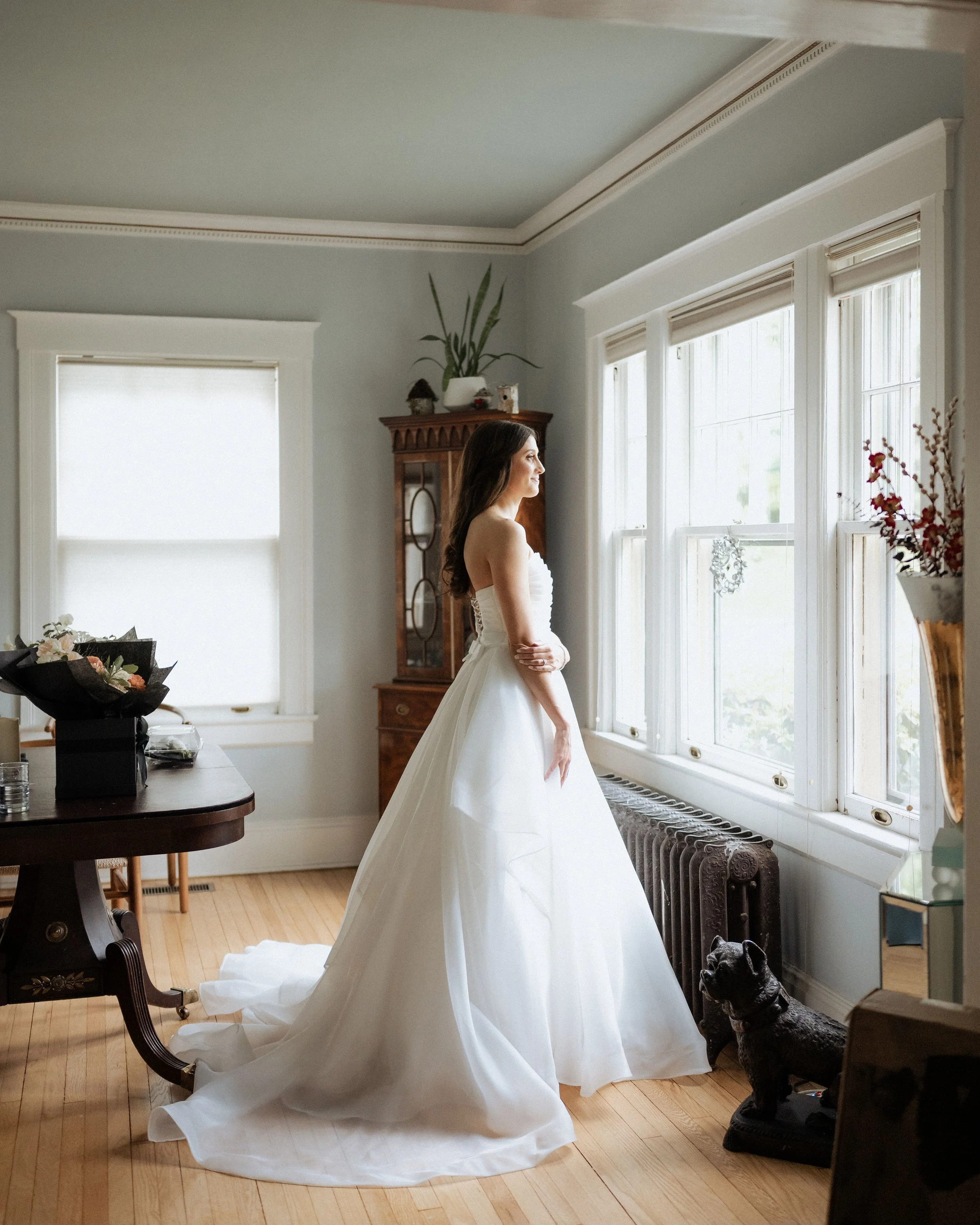 A woman in a wedding dress standing by a large window, looking outside in a well-lit room with wooden furniture and a small black dog nearby.