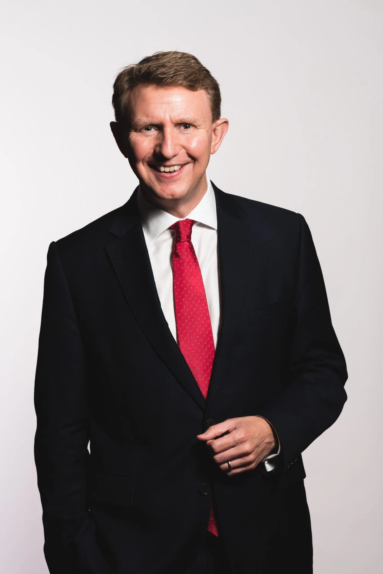 A smiling man in a black suit, white shirt, and red tie, standing against a plain white background.