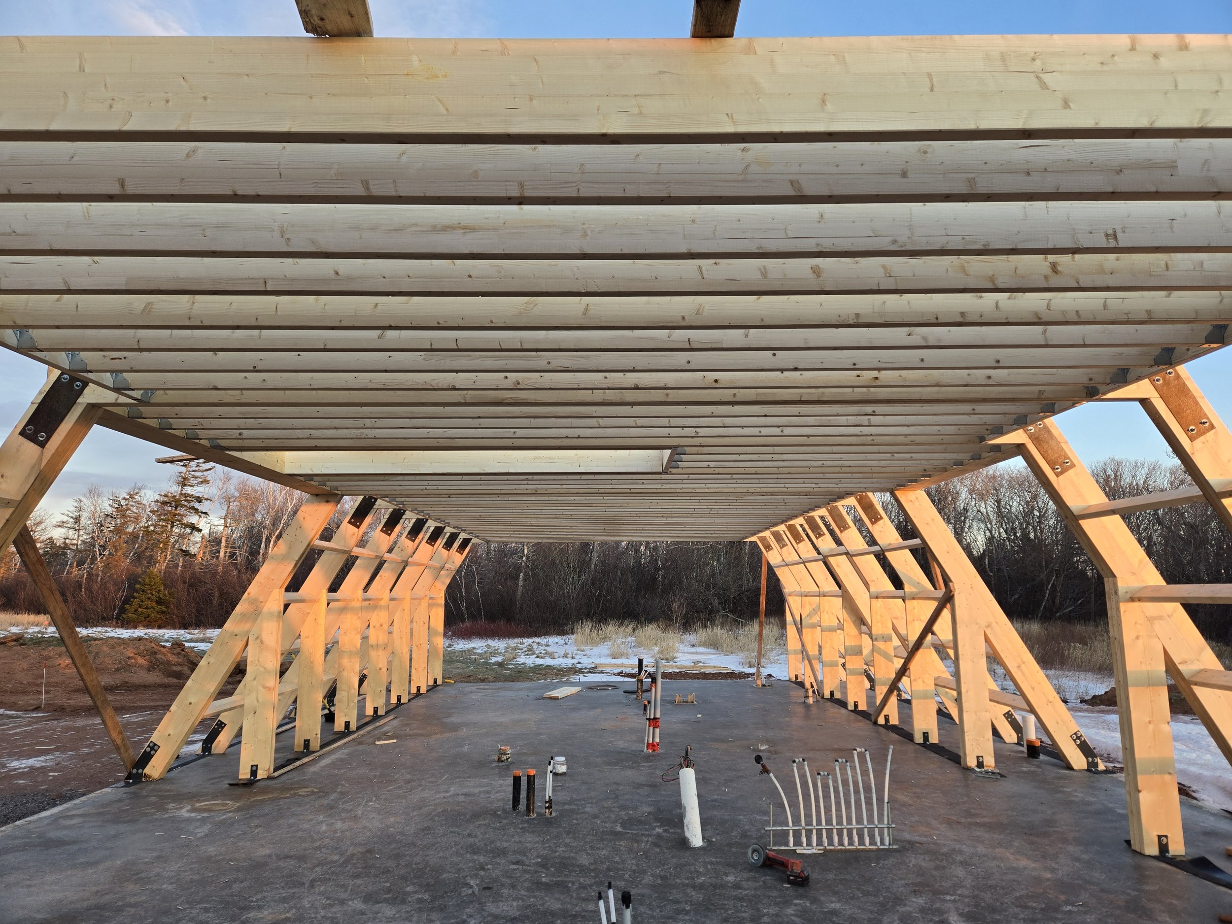 Under construction wooden building framework with wooden beams and support structures, outdoor setting with trees and clear sky.