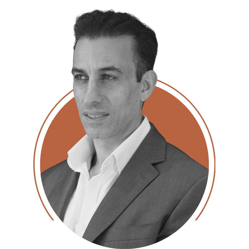 Black and white professional headshot of Andonis Petrou, Founder + CEO of Teclab Ltd, a man with dark neatly styled hair, wearing a dark suit and white dress shirt, against a solid background with an orange circular border.
