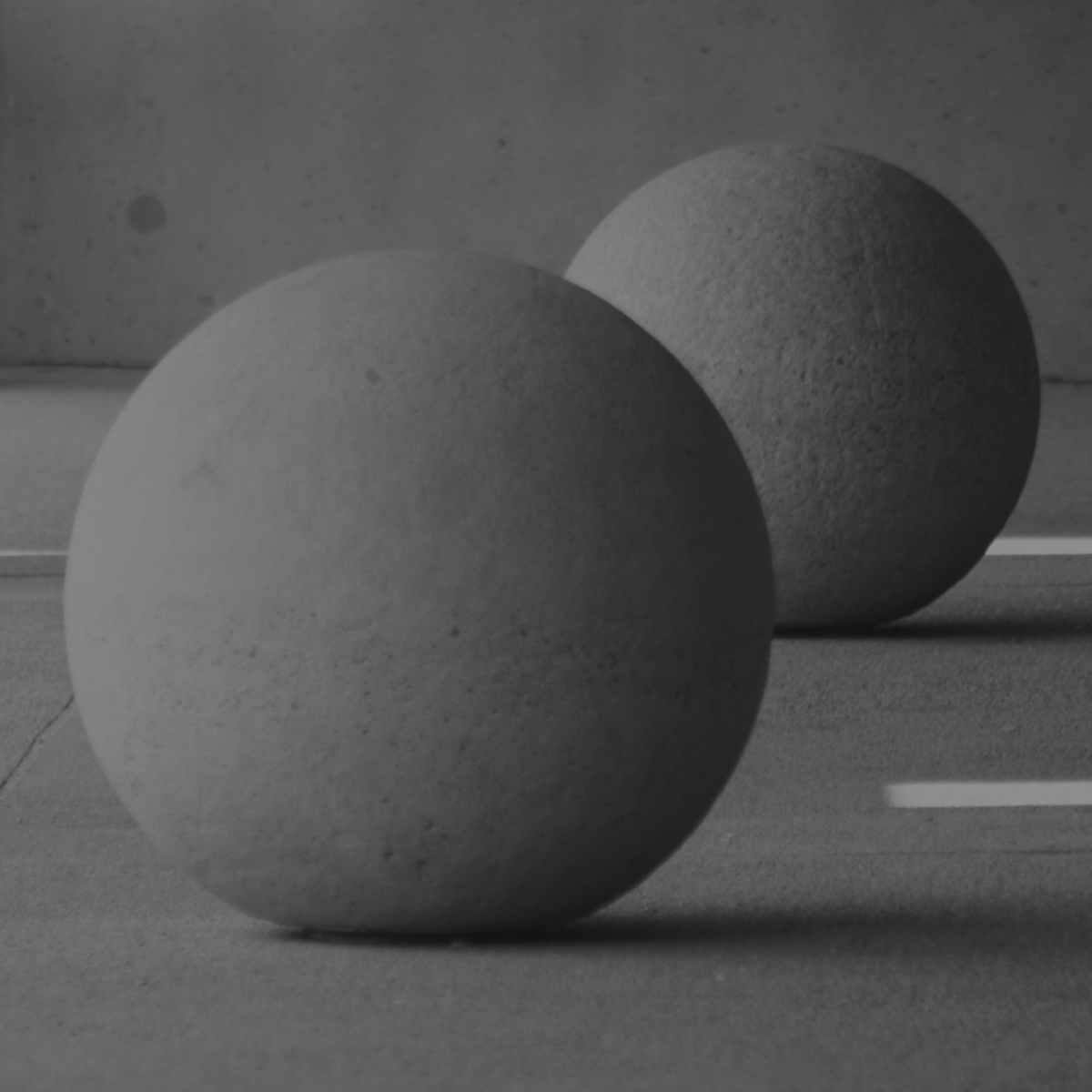 Two concrete balls placed on a paved surface against a concrete wall, with shadows casting on the ground.
