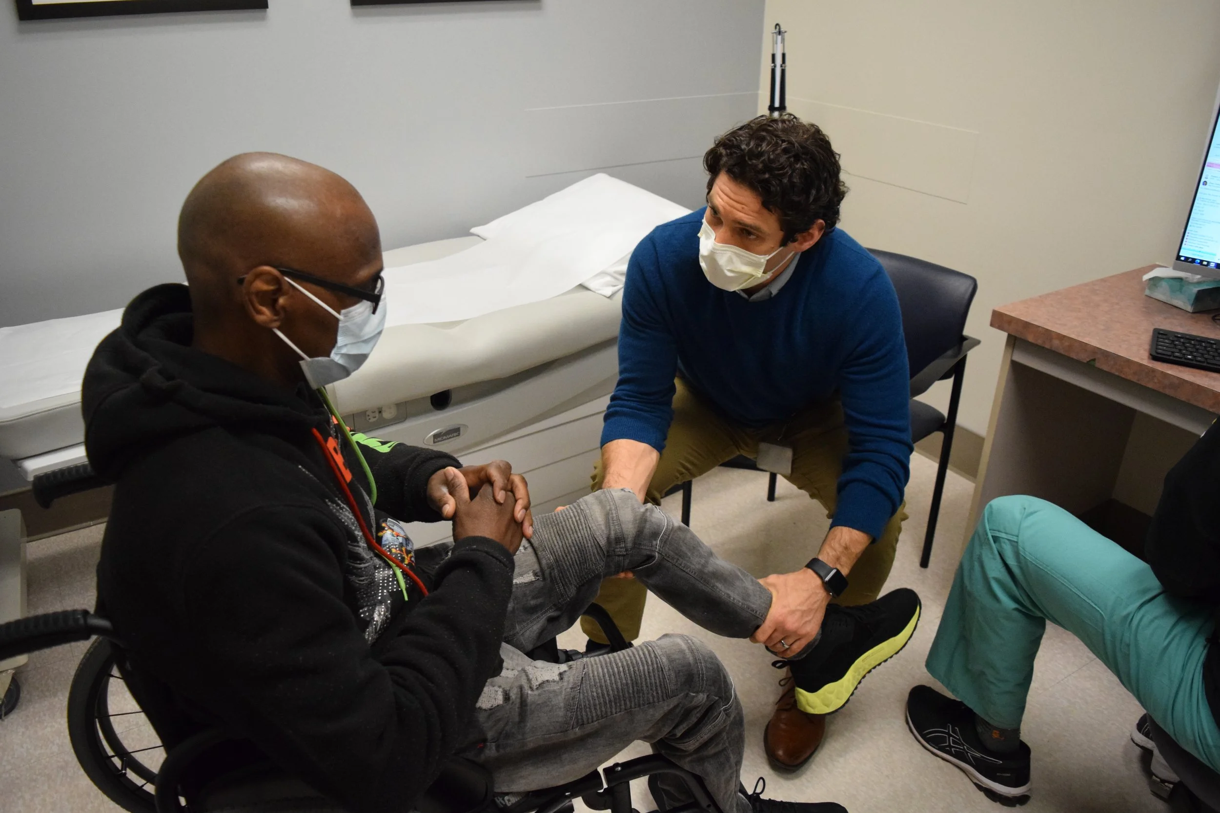 Dr. Neudorf sitting in front a male patient sitting in a wheelchair, holding and examining his leg. The patient in the wheelchair looks down at his leg. Both are wearing masks in a hospital room with medical bed and equipment.