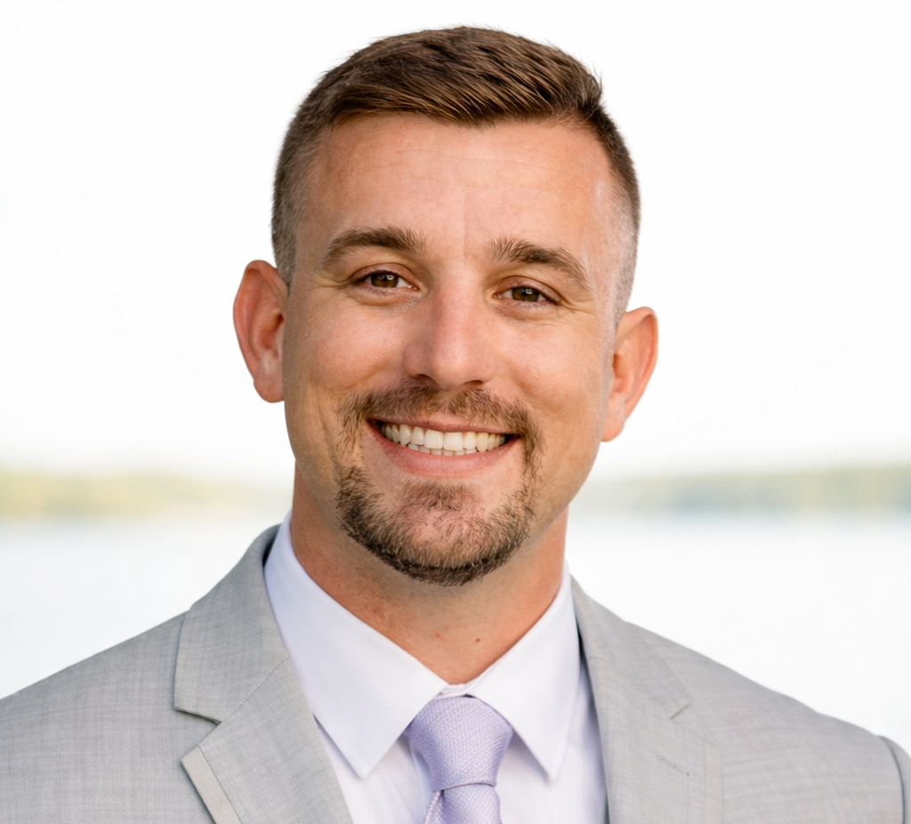 A smiling man in a light gray suit and tie poses outdoors against a blurred background of water and sky.