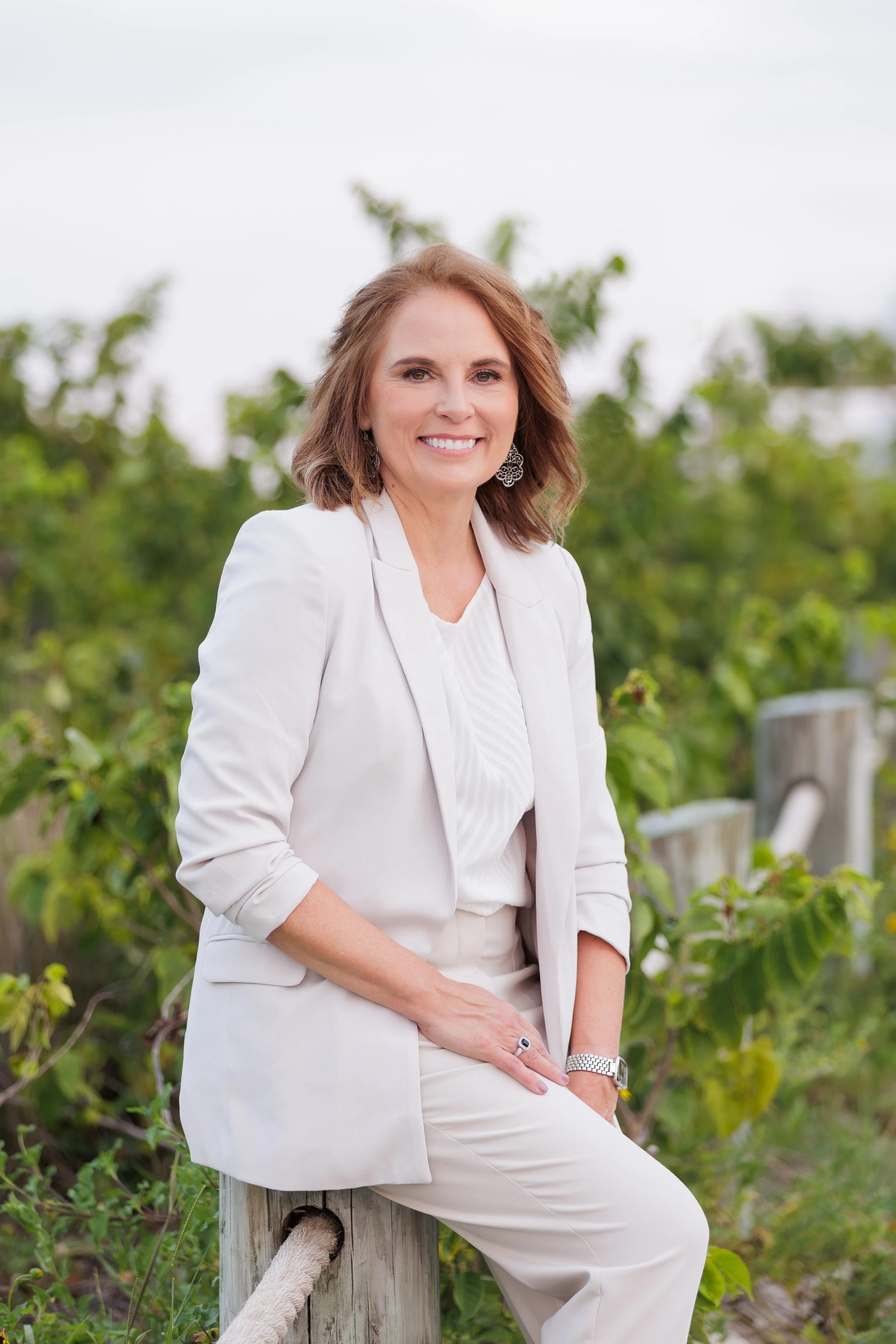 A woman with shoulder-length brown hair smiling, dressed in a white blazer and pants, sitting on a wooden post outdoors with green foliage in the background.
