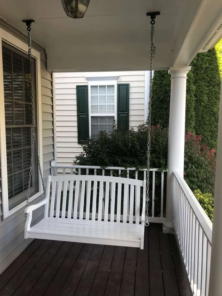 Front porch with a white wooden swing hanging from the ceiling, adjacent to white siding house with a window, surrounded by greenery and bushes.