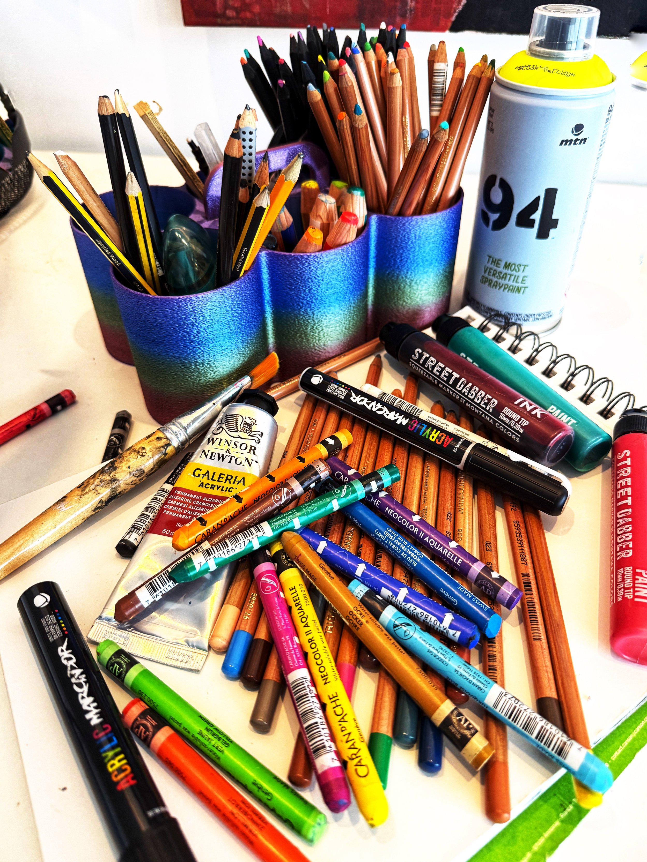 Mixed media tools laid out on a table including a sketch pad, water soluble crayons, paint markrs, acyrlic paints, brushes, colored pencils, and woody stabilos