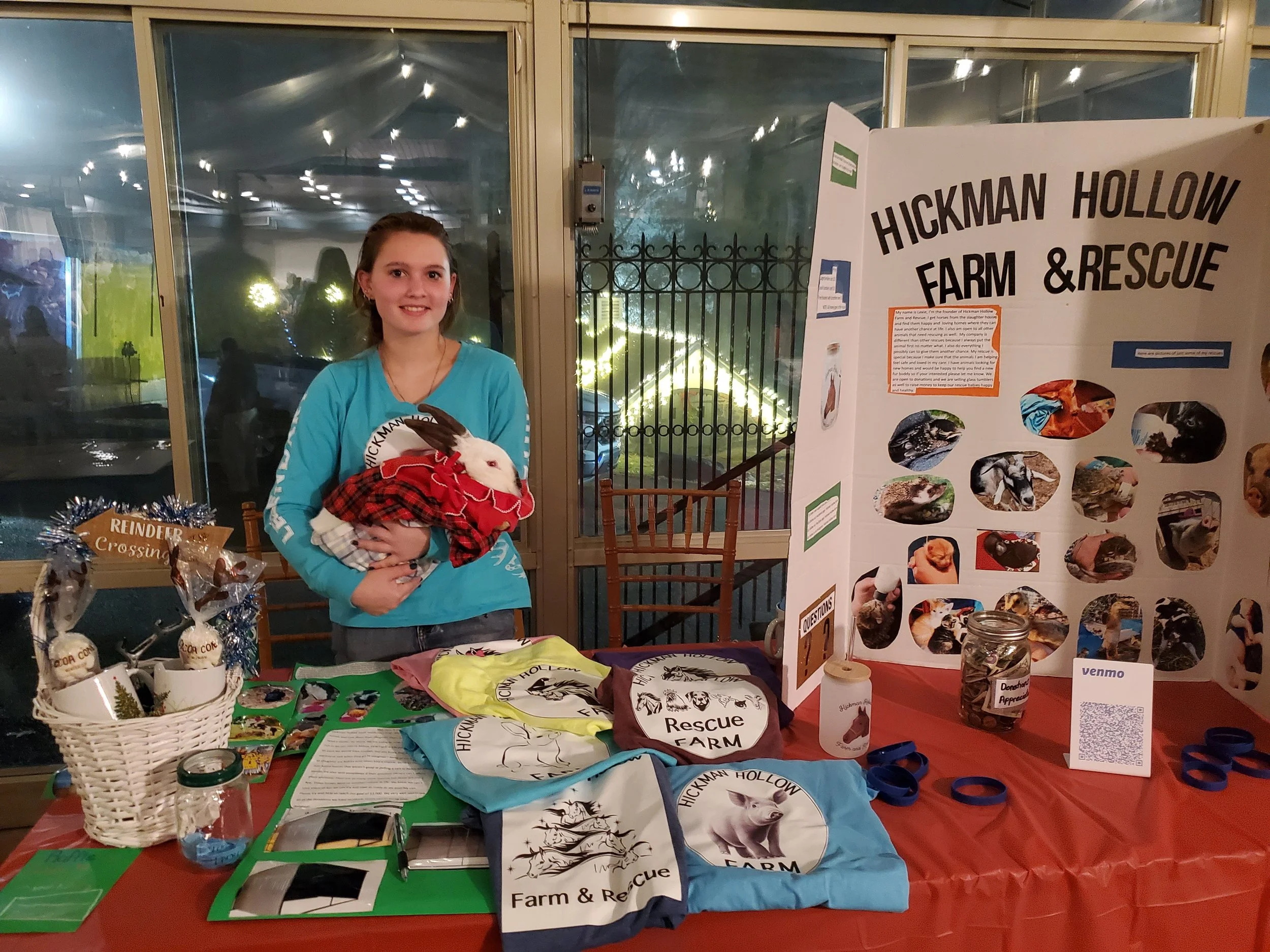 A young woman holding a white rabbit wearing a red and black plaid outfit at an event booth for Hickman Hollow Farm & Rescue, with informational posters, merchandise, and donation jars on the table.