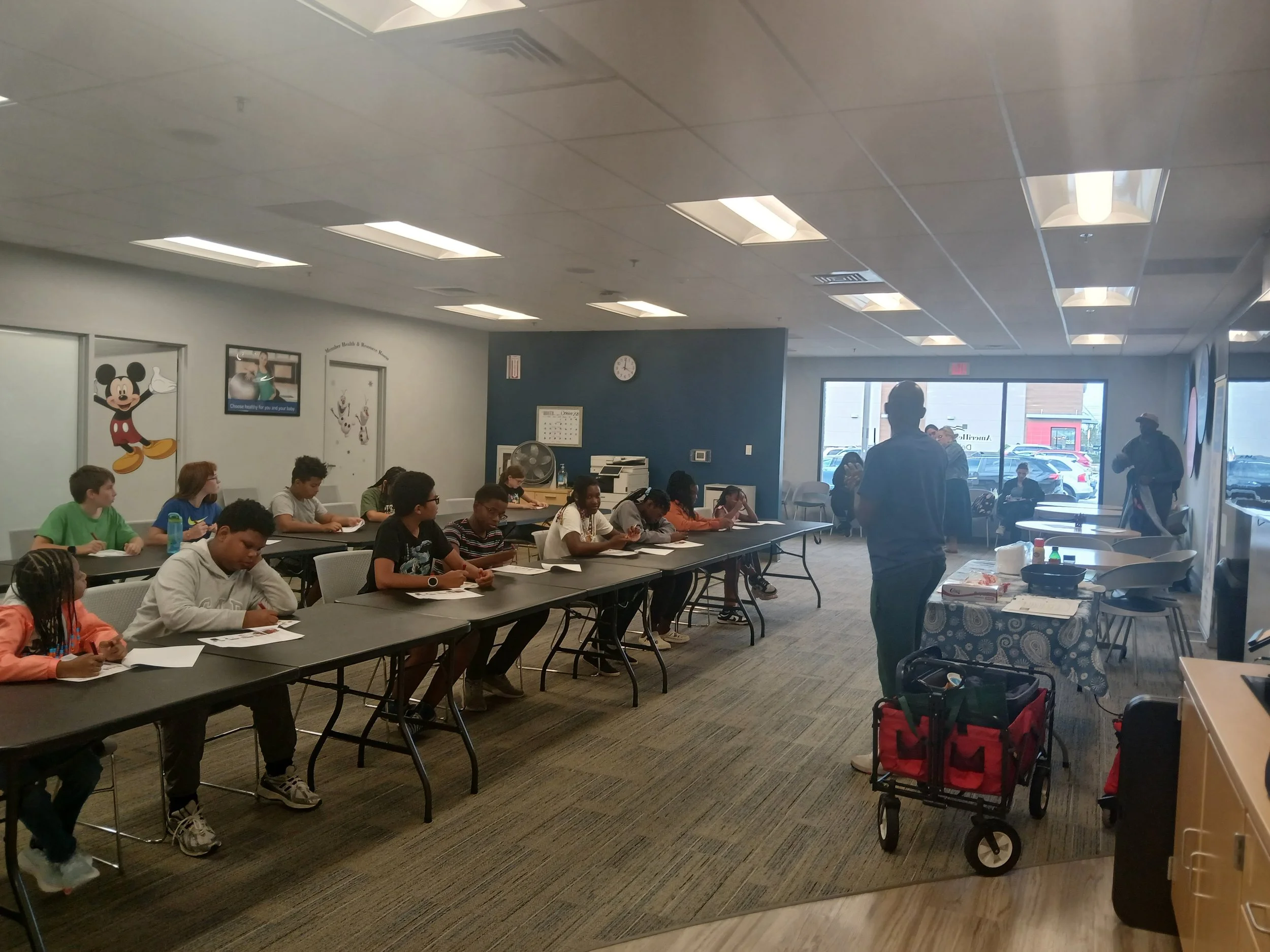 Children sitting at tables in a classroom or childcare center with Mickey Mouse decoration on the wall, instructors standing at the front, and a table with supplies.