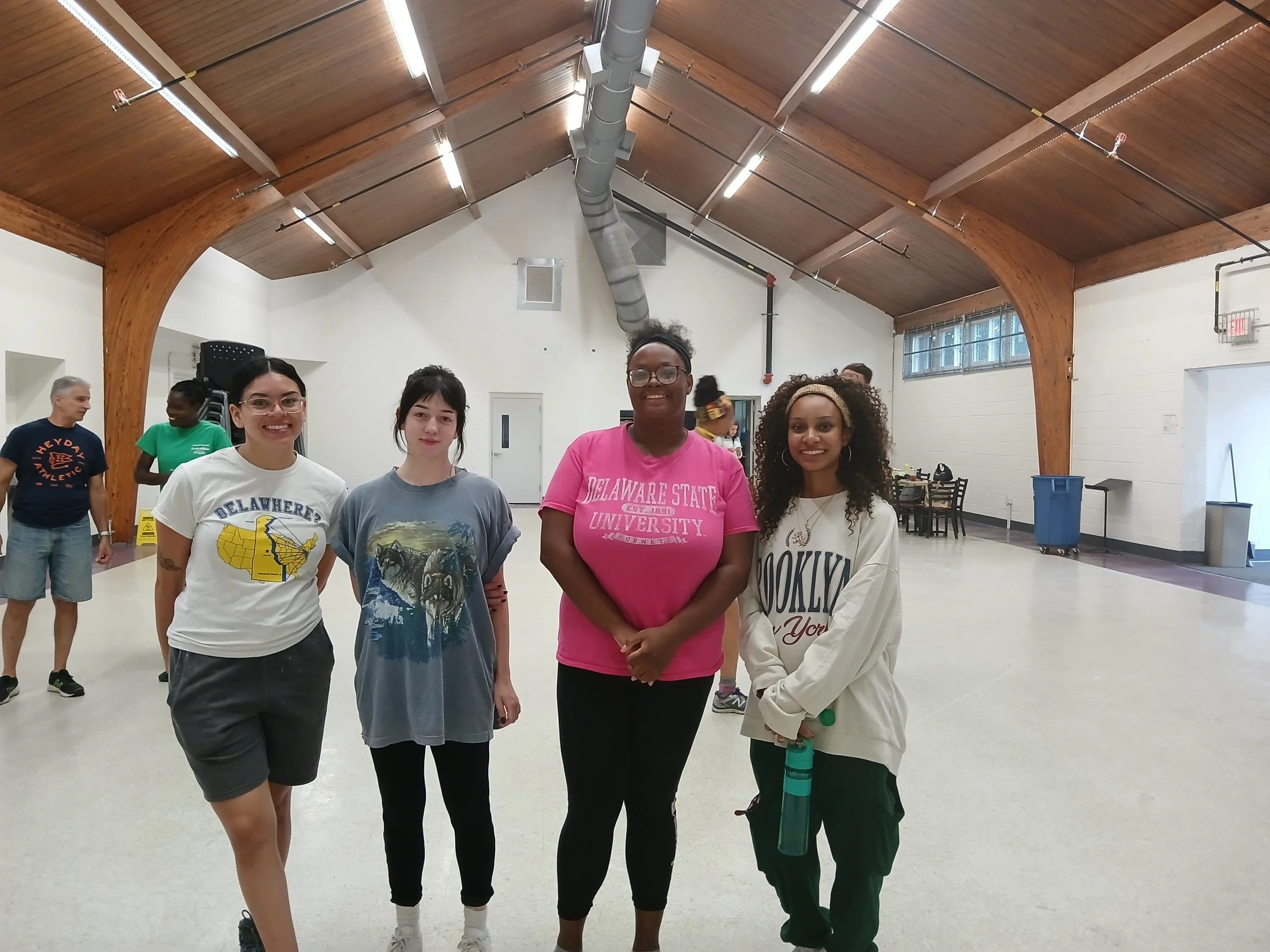 Four women standing together in a large indoor space with wooden beams on the ceiling. They are smiling at the camera, with people and tables visible in the background.