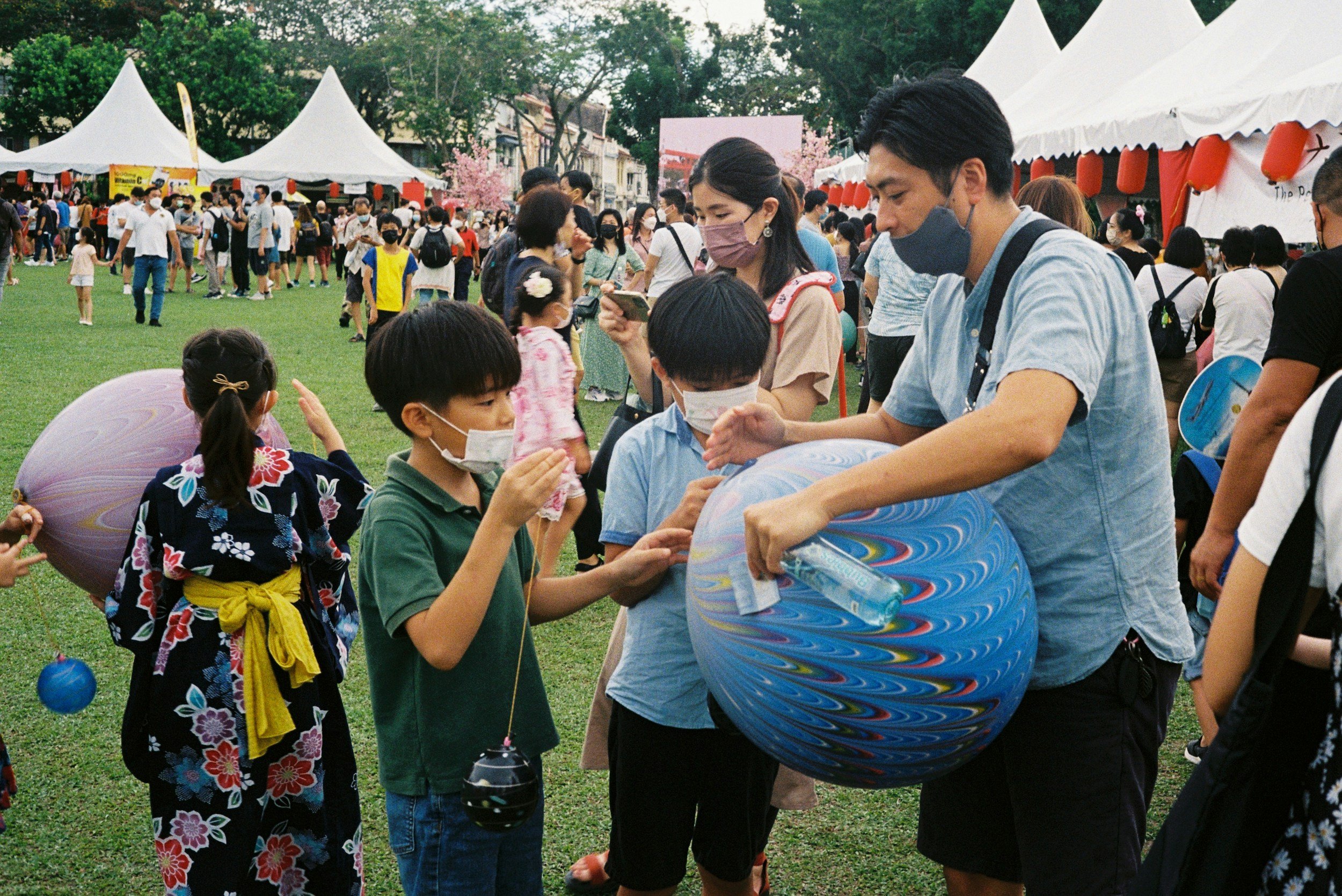 People at an outdoor festival with tents, children, and adults, some wearing masks. Children are holding lanterns and talking to a man holding a painted paper lantern.