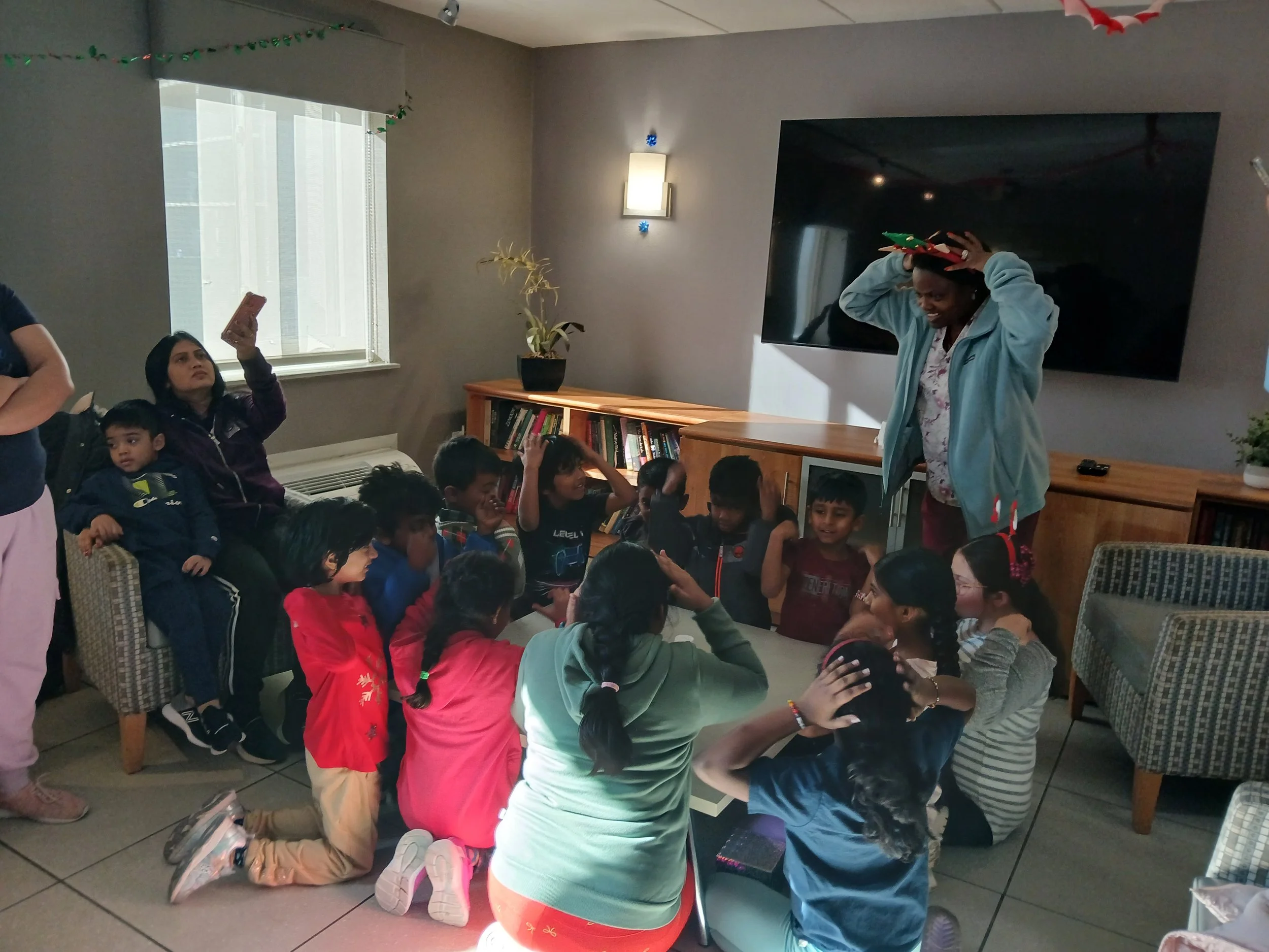 A group of children and adults gather in a room with a wall-mounted TV, decorated for a celebration, with one person wearing a festive hat. One woman is sitting on a chair taking a selfie, and the others are sitting or kneeling around a low table, engaging in an activity.