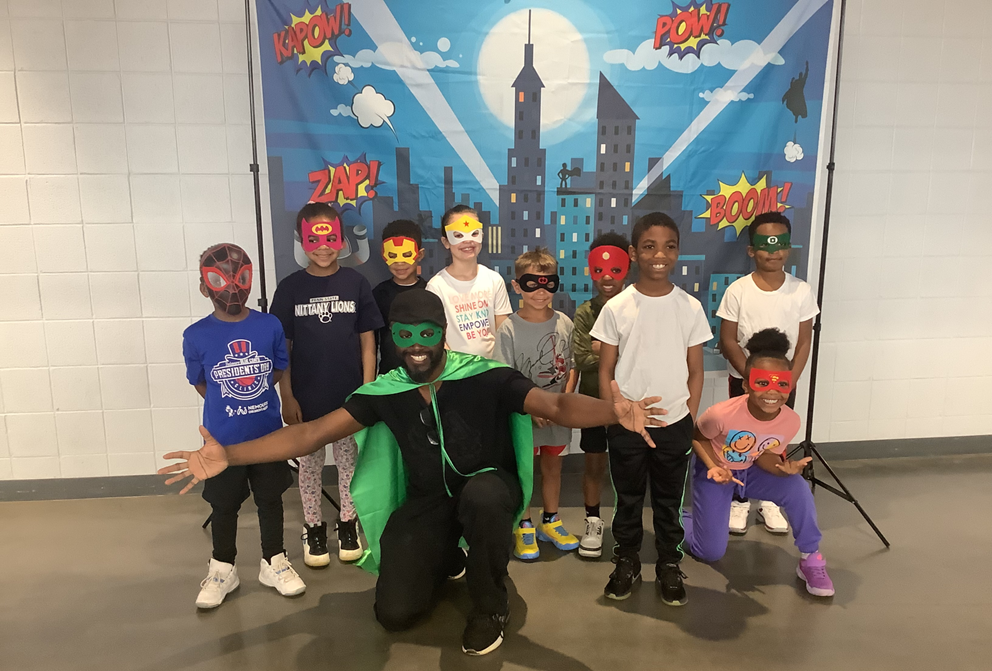 Group of children and an adult dressed as a superhero, all wearing colorful superhero masks, in front of a comic book style backdrop with city buildings and words like 'Kapow!', 'Zap!', 'Pow!', and 'Boom!'.