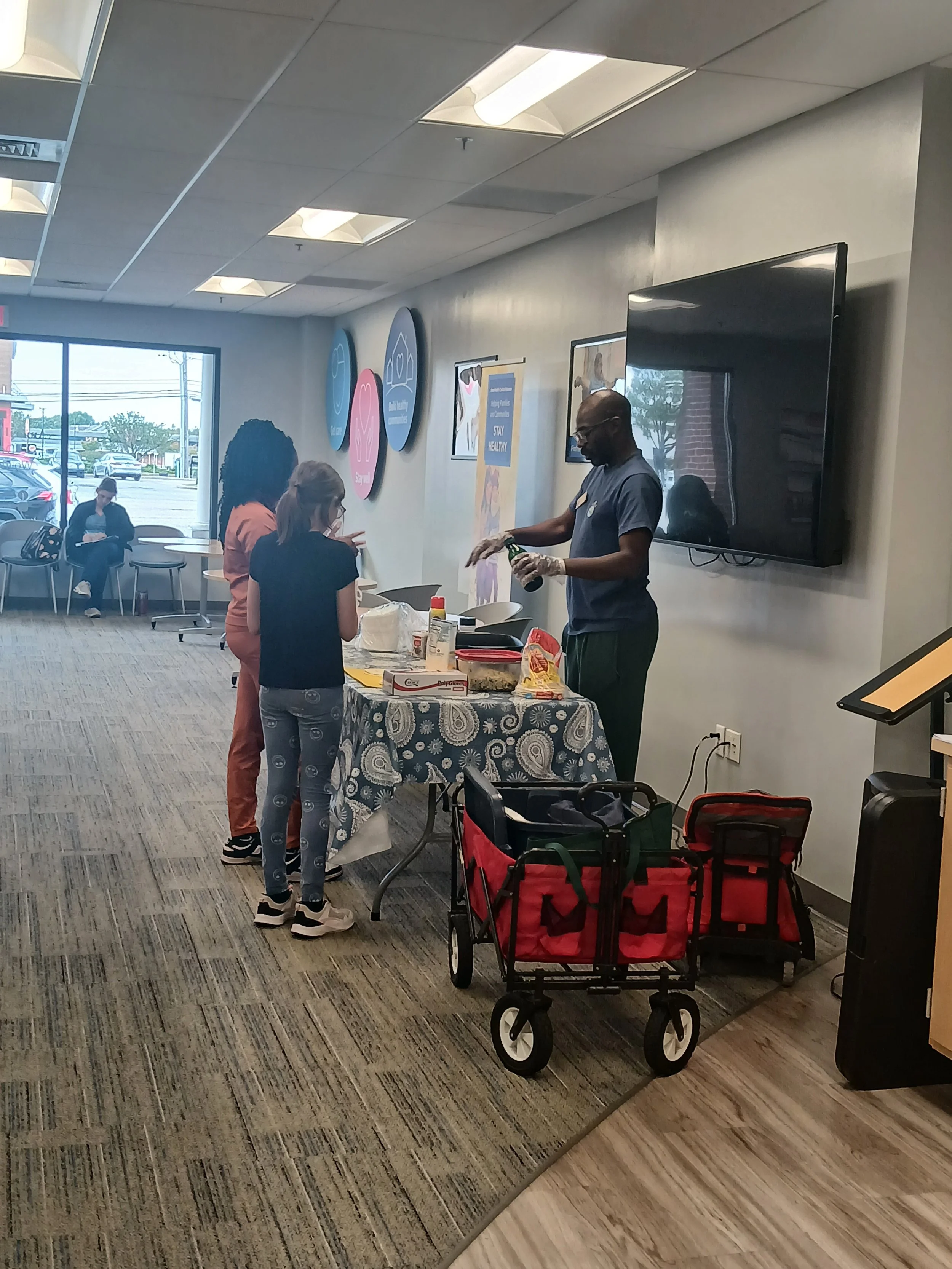 A man preparing food at a table with children in line, inside a room with large windows and a TV on the wall.