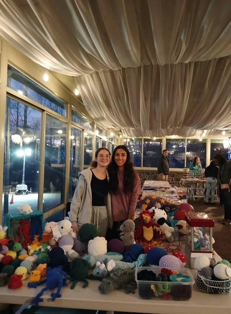 Two women smiling at a table filled with colorful handmade knitted and crochet stuffed animals at an indoor market or craft fair.