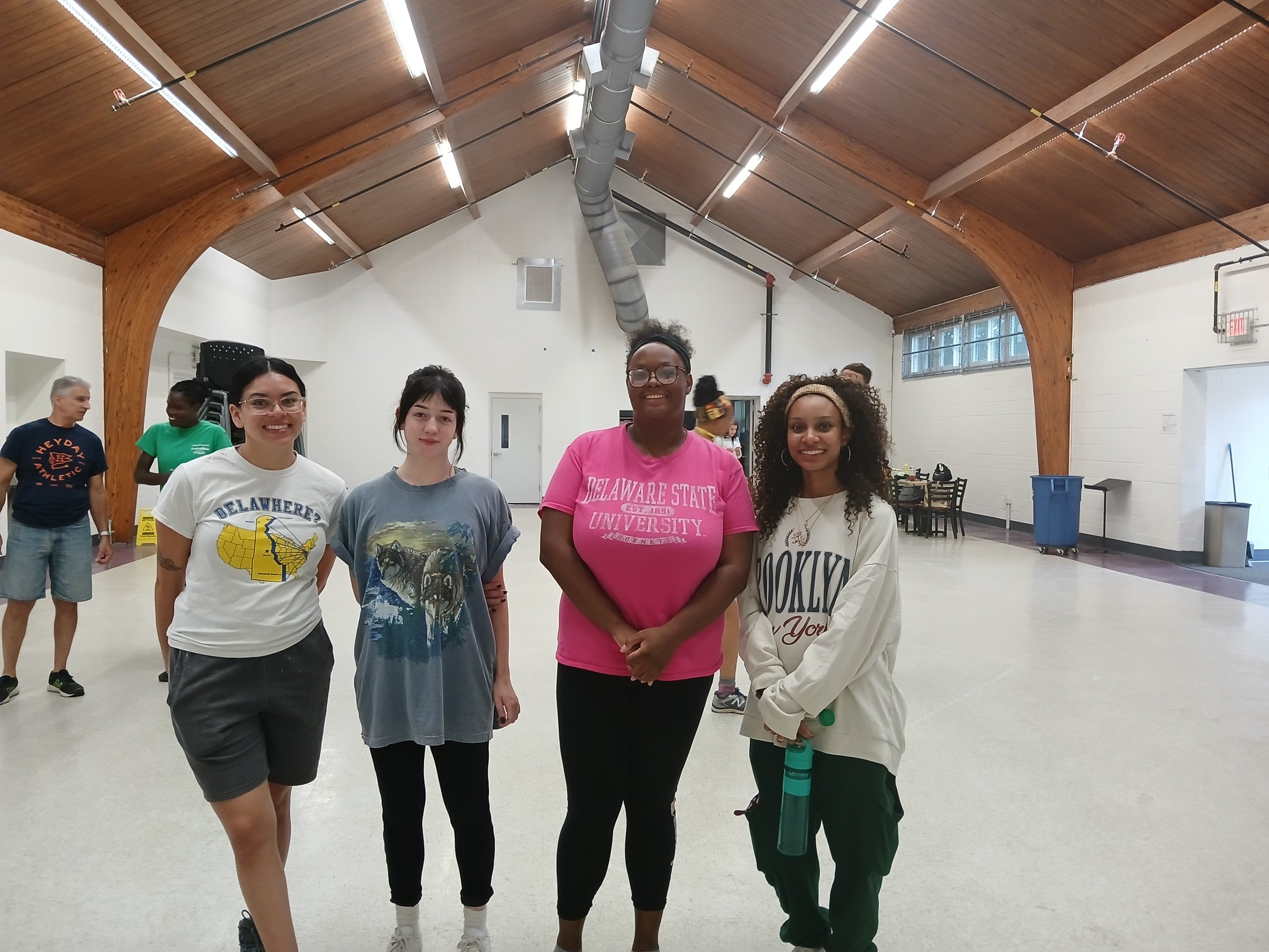 Four women standing together in a gymnasium, smiling at the camera. In the background, two people are walking and talking, with gym equipment and a wooden ceiling visible.