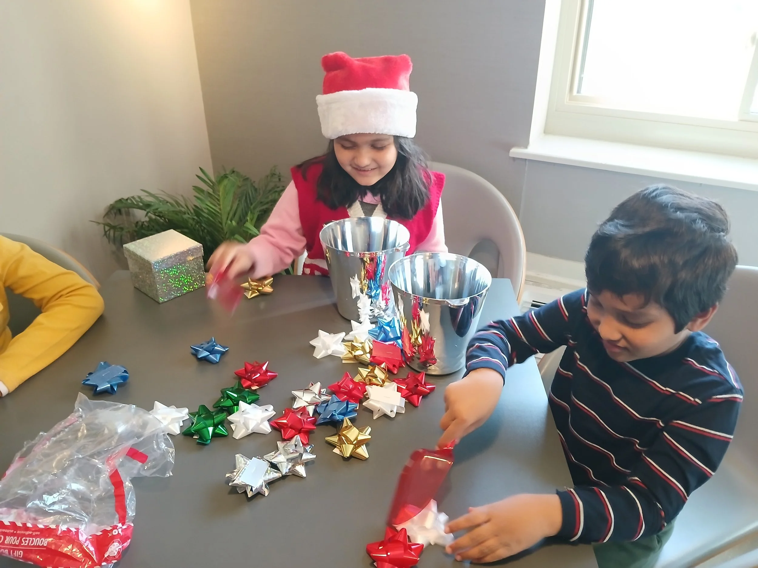 Two children sitting at a table, wrapping Christmas presents with colorful bows, one girl wearing a Santa hat and a red vest, the other boy in a striped shirt, with two metal buckets and a small silver gift box on the table.