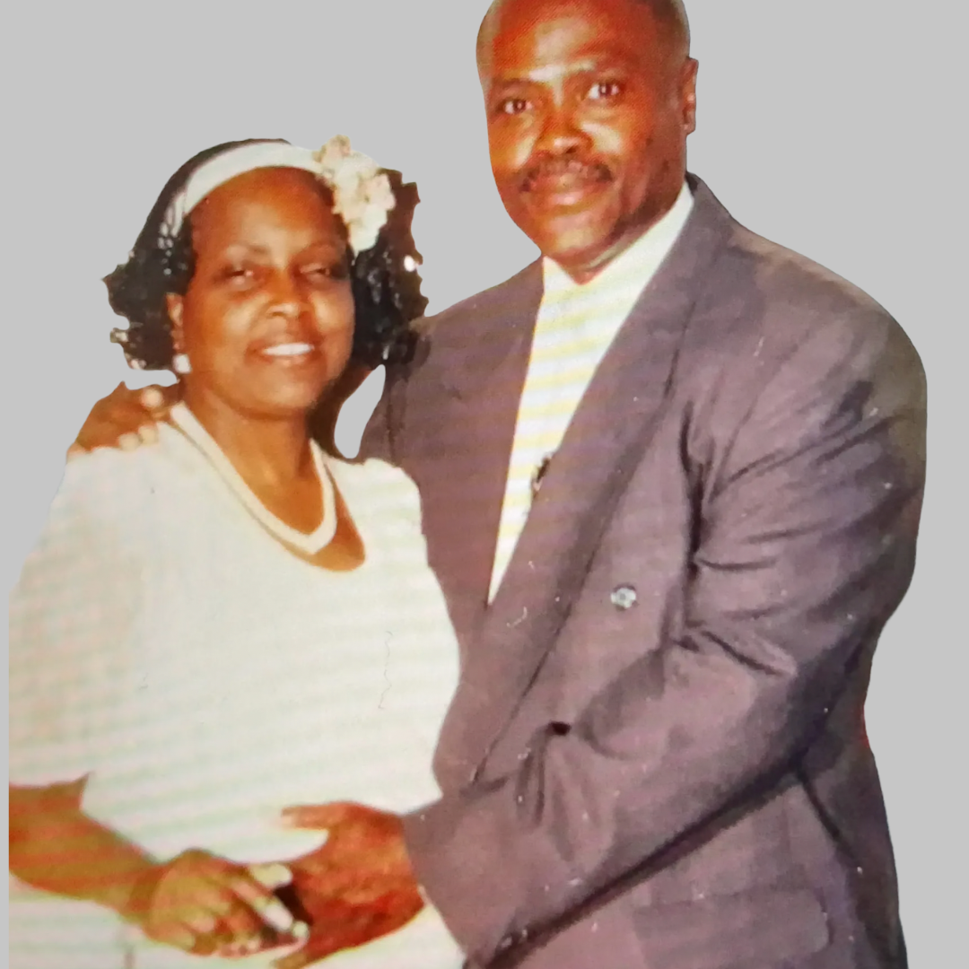 A man and woman standing close together, smiling, with their hands clasped. The woman has curly hair with a headband and wears a white outfit. The man has a mustache and wears a dark suit with a striped shirt.