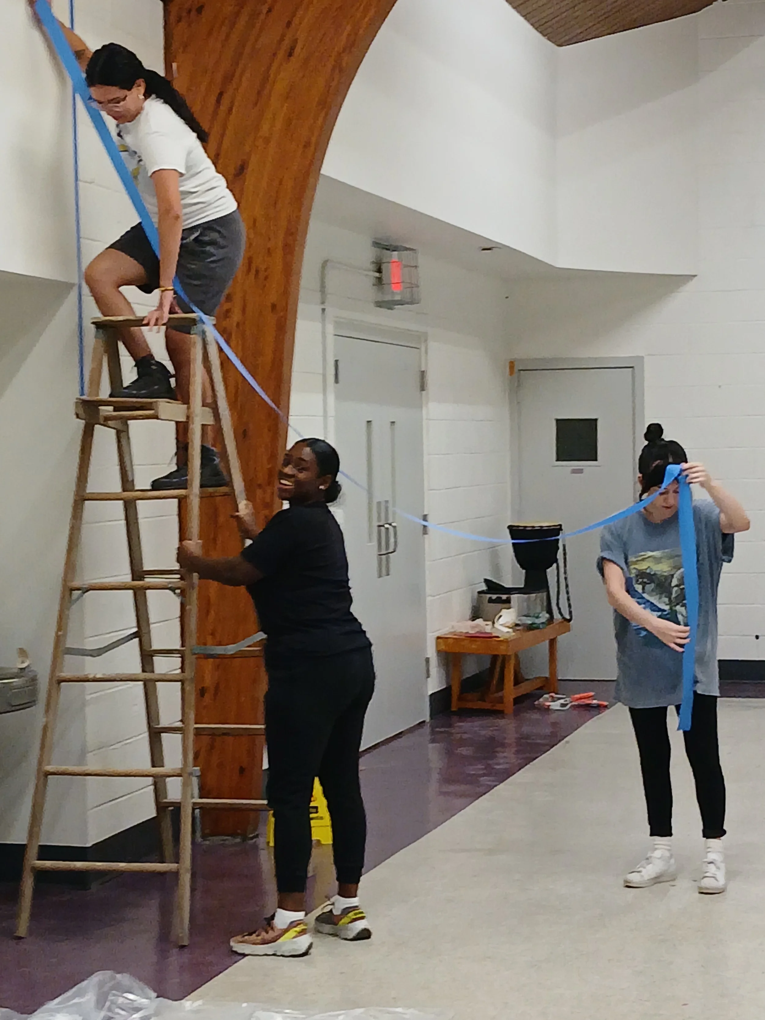 Three women are hanging blue ribbons on a wooden ladder, with one woman sitting on top and two women holding the ladder while standing on the ground.