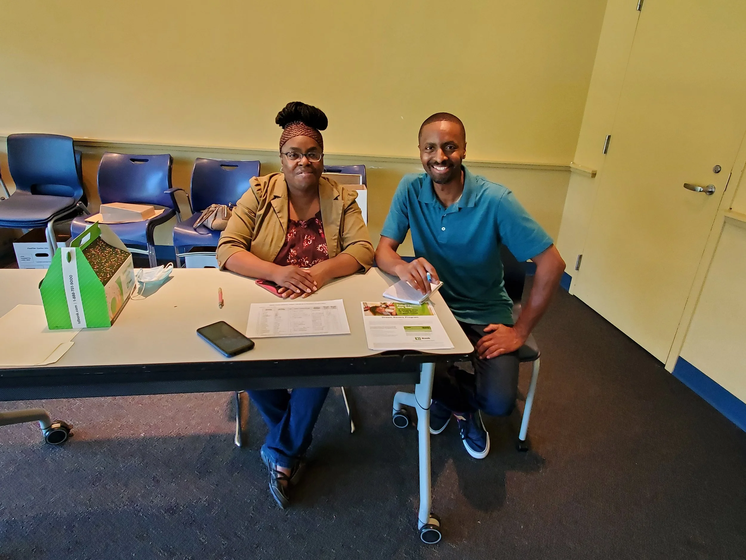 Two people seated at a table with documents, a pen, a smartphone, and a box of tissues, in a room with chairs against a yellow wall.