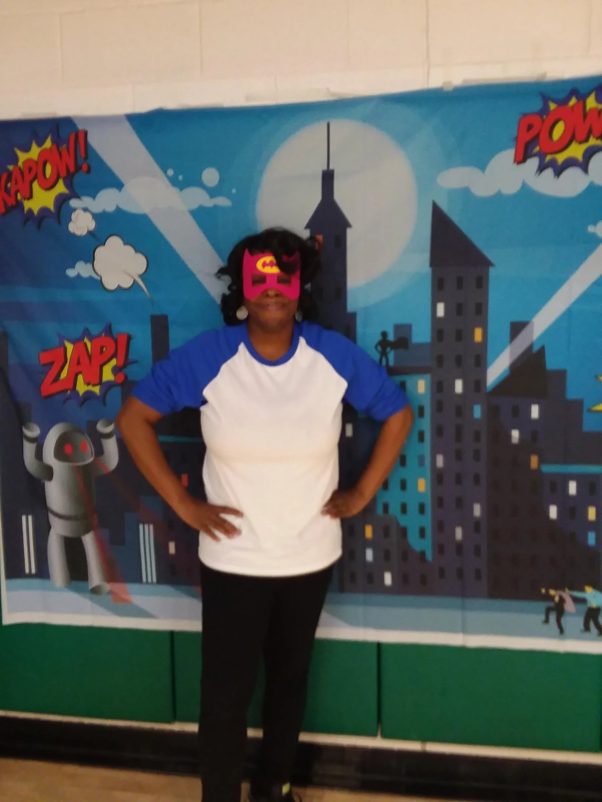 Person wearing a pink superhero mask with a Batman logo, white and blue shirt, standing in front of a comic book style cityscape backdrop with words like 'KAPOW!', 'ZAP!', and 'POW!'.