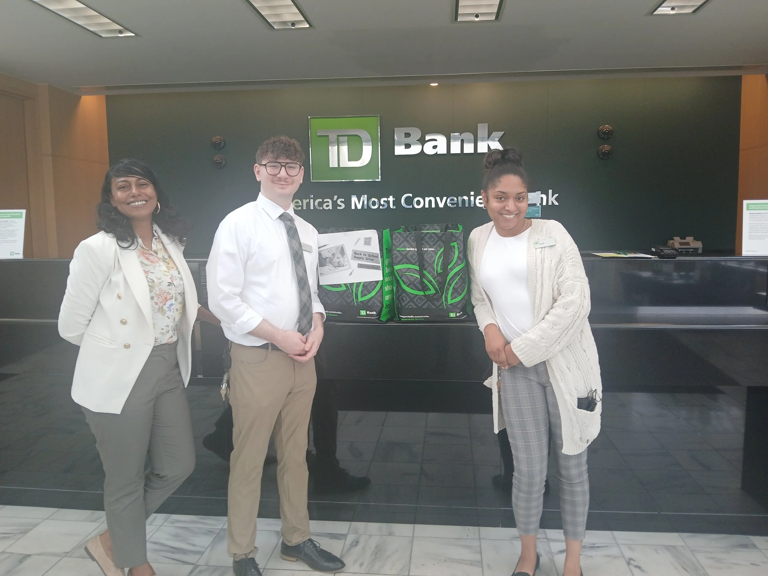 Three people standing behind a counter at a TD Bank branch, smiling for a photo. The background shows the bank’s logo and slogan, with bags on the counter in front of them.