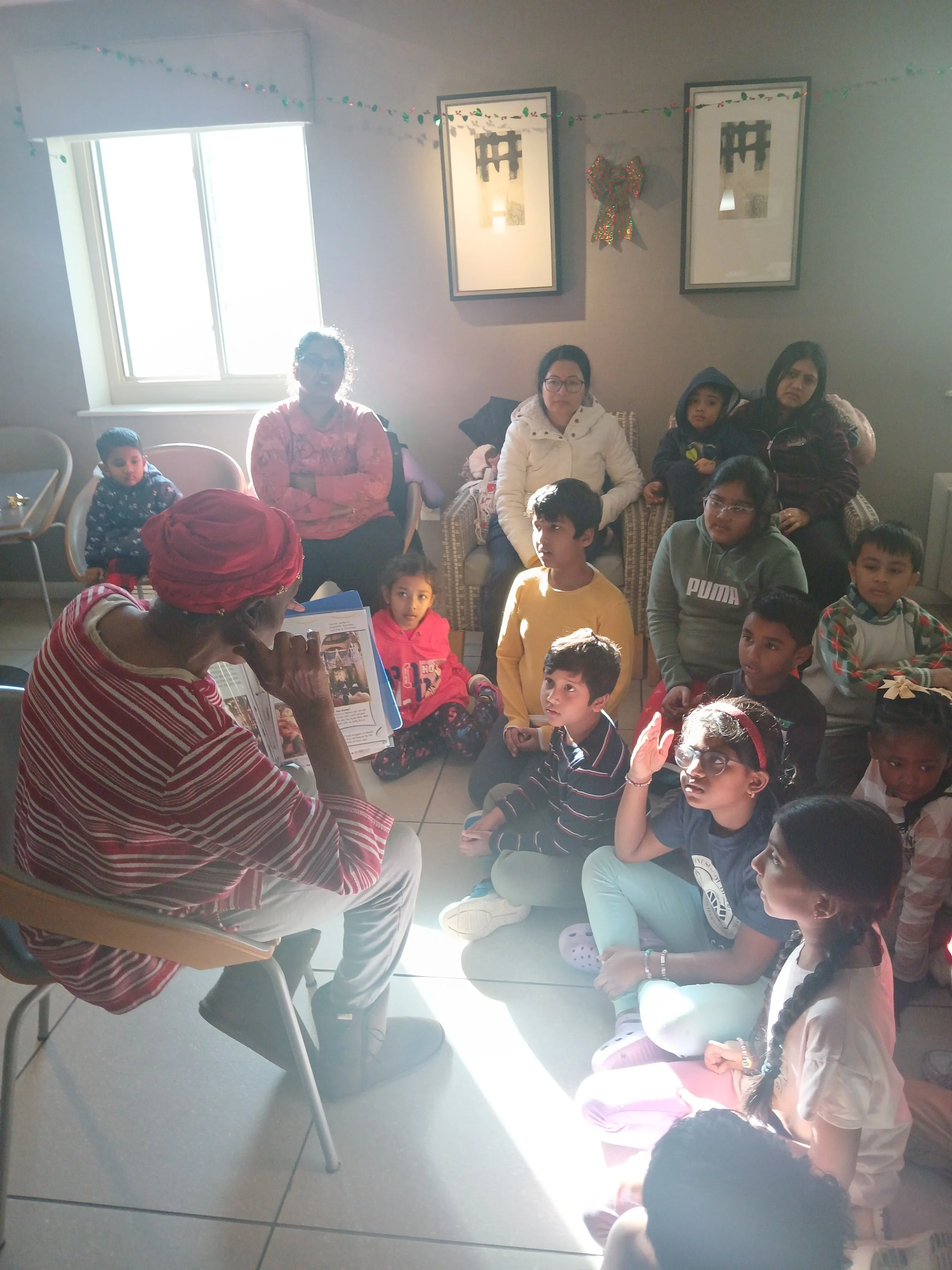 An elderly person wearing a red turban and striped shirt reading a book to a group of children and adults seated on the floor and chairs in a brightly lit room.