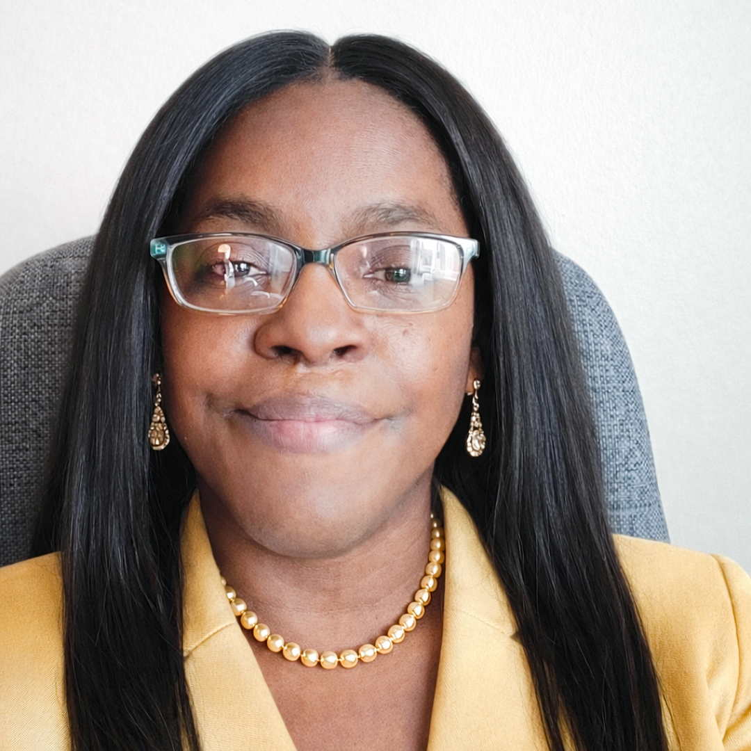 A woman with dark hair, wearing glasses, pearl earrings, and a pearl necklace, sitting in a gray chair, wearing a yellow blazer, smiling in front of a light-colored wall.