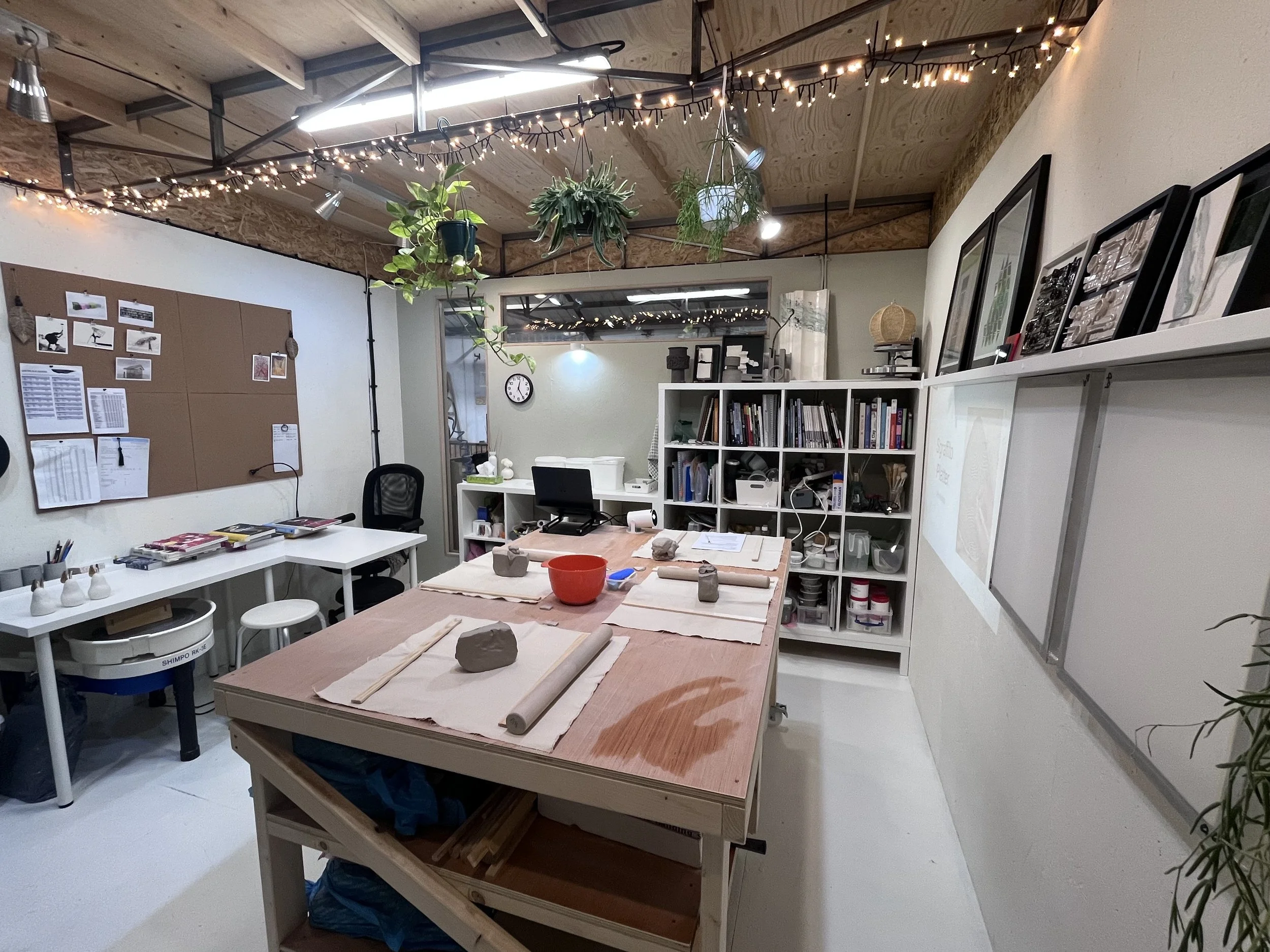 A well-lit art studio with a large wooden worktable in the center, covered with clay and tools. Shelves filled with art supplies, framed artwork, and books line the walls. Potted plants hang from the ceiling, and string lights add a cozy atmosphere. A whiteboard is on the right wall, and a corkboard with papers and photos hangs on the left wall.