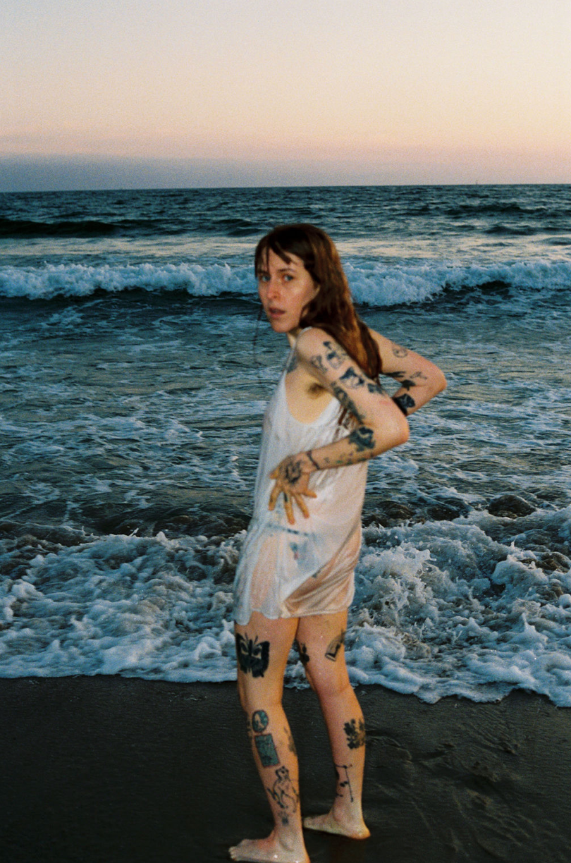 A young woman with tattoos on her arms and legs stands barefoot in the surf of a beach at sunset, looking back over her shoulder.