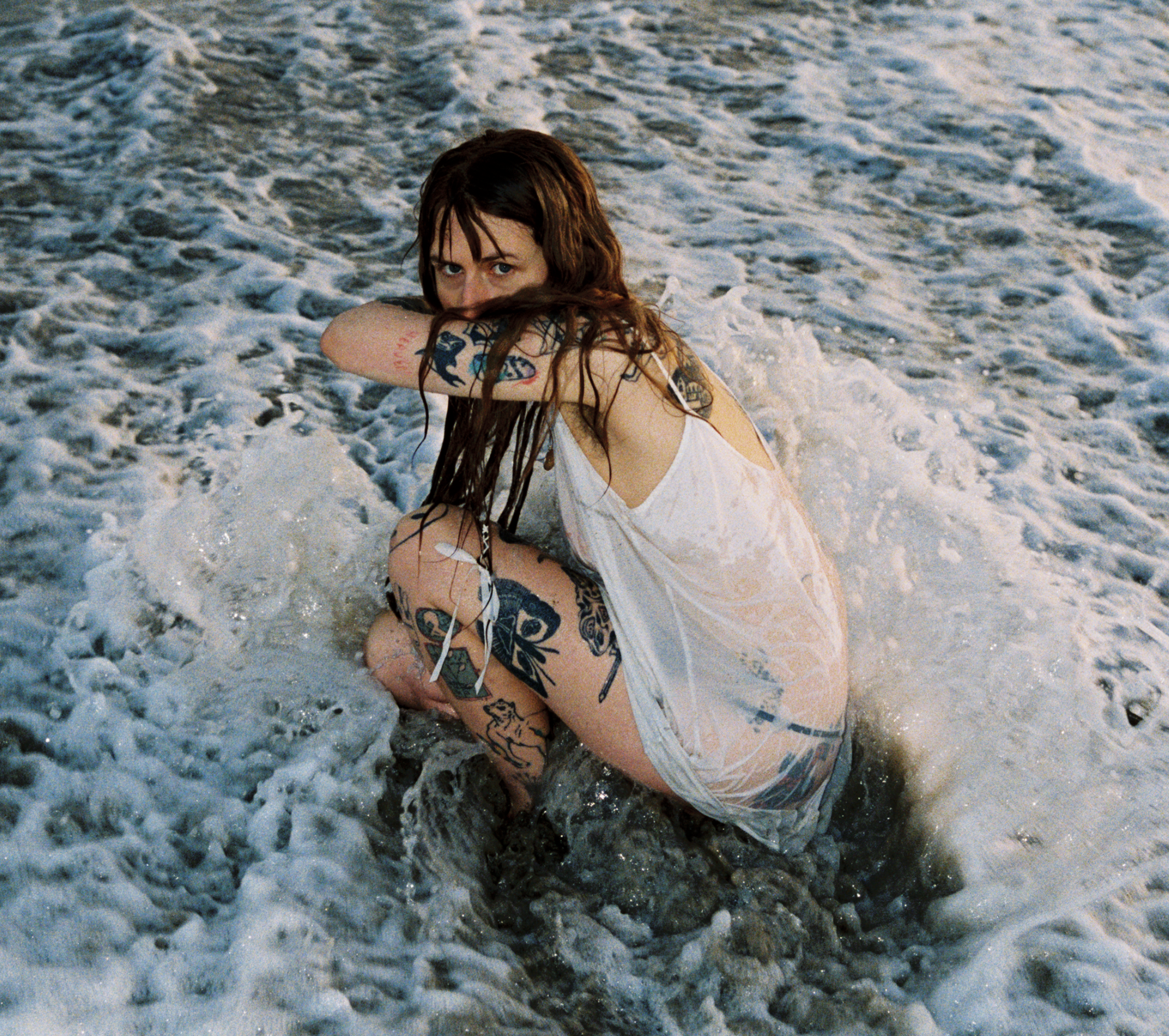 Woman with tattoos squatting in ocean waves, wearing a wet white dress, looking at the camera.