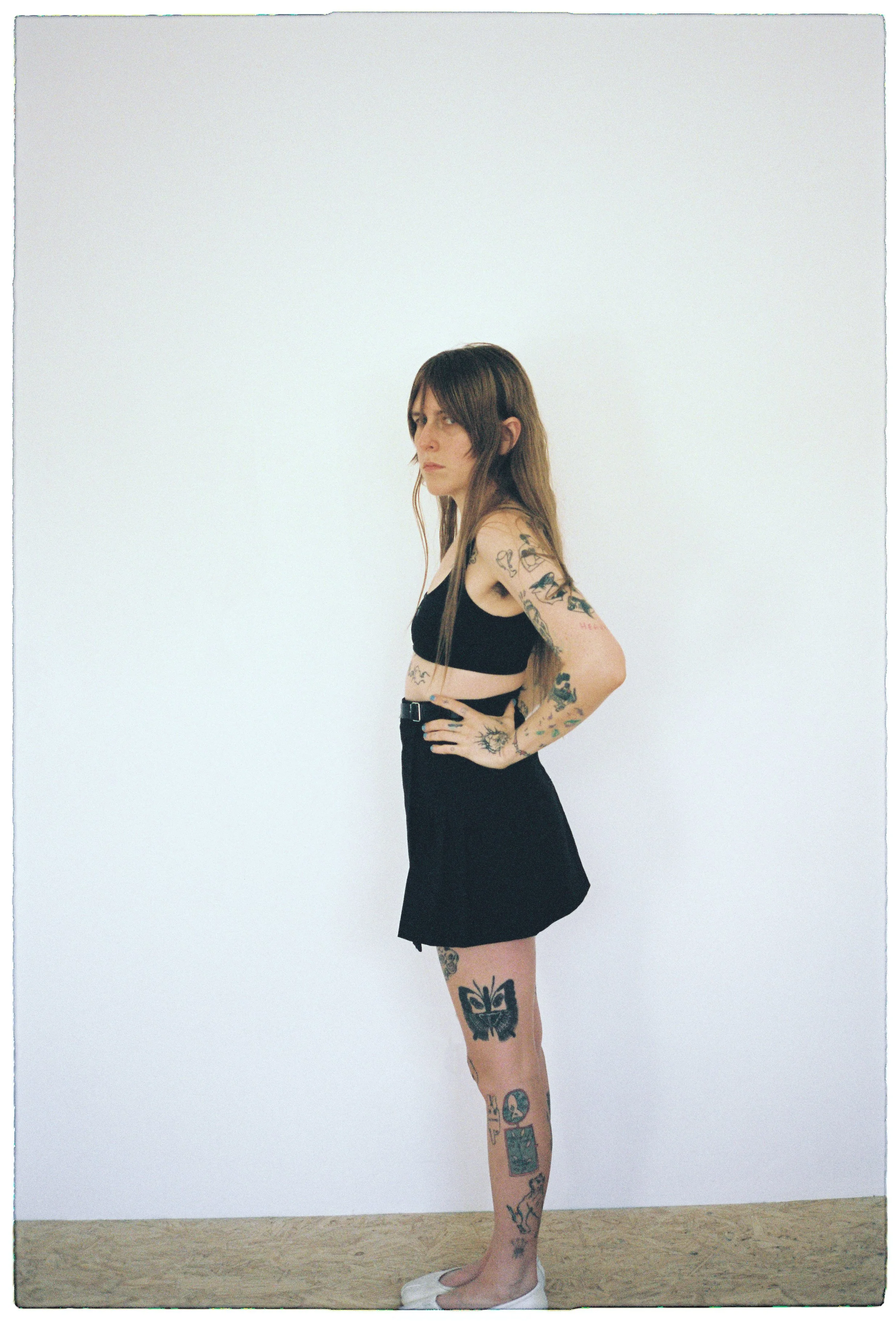 A young woman with long brown hair and multiple tattoos stands in profile against a plain white wall. She wears a black crop top, black skirt, and white shoes, with her hands resting on her hips.
