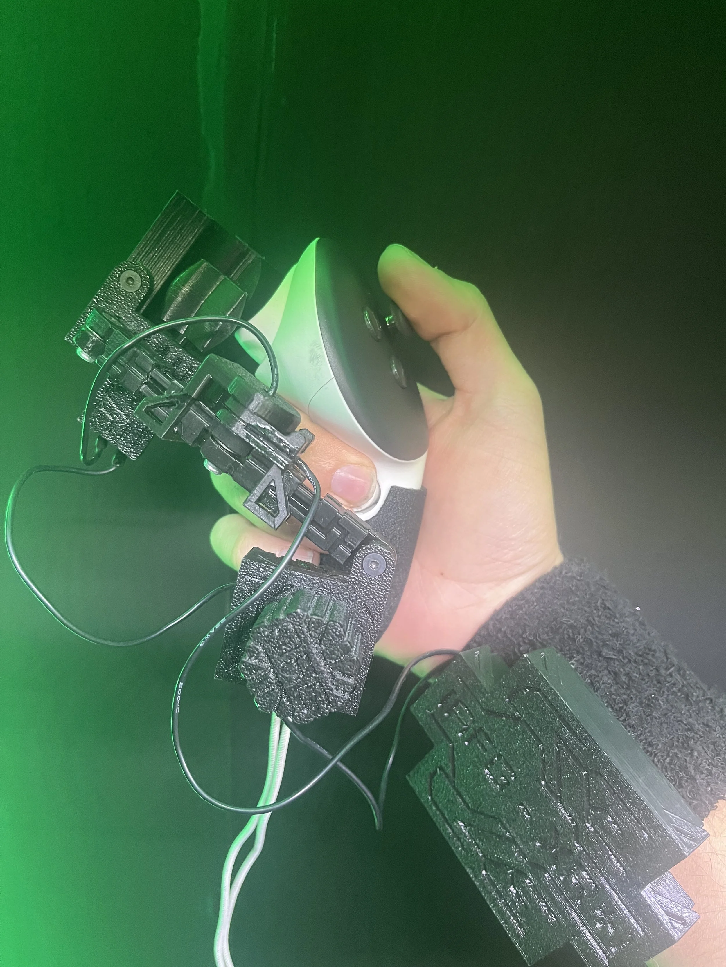 Hand holding a small electronic device with wires, foam padding, and a circuit board attached, against a dark background with green light.