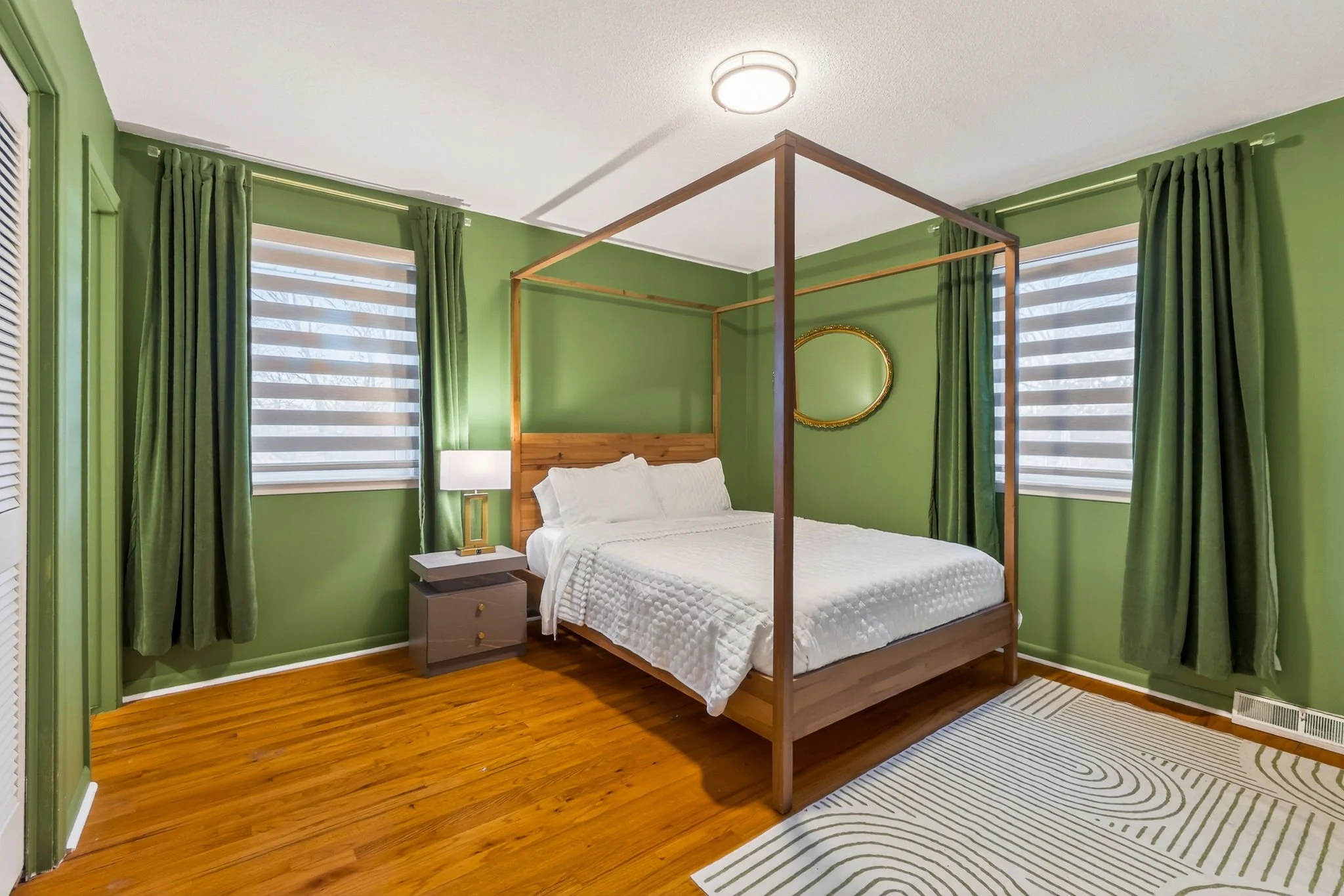 A bedroom with bamboo floor, green walls, and two large windows with green curtains and horizontal blinds. There's a wooden four-poster bed with white bedding, a small bedside table with a lamp, a gold oval mirror on the wall, and a striped area rug 