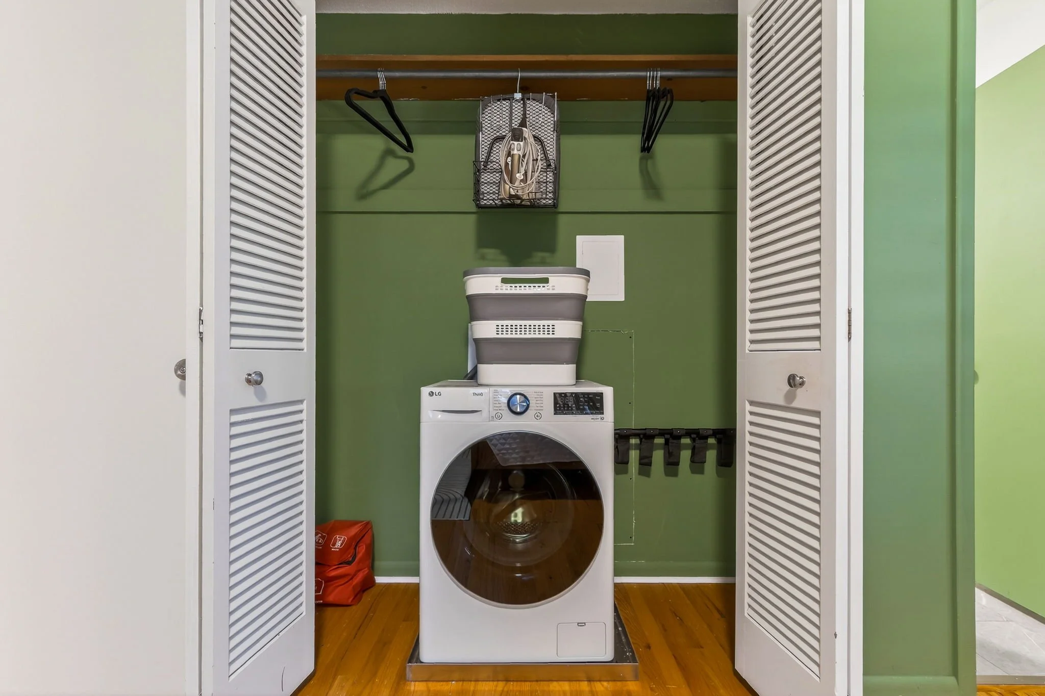 Laundry closet with open white louvered doors, containing an LG front-loading washing machine. On top, there are cleaning supplies stacked, and above, a shelf with hangers and a basket holding an iron. The back wall is painted green, and the floor is