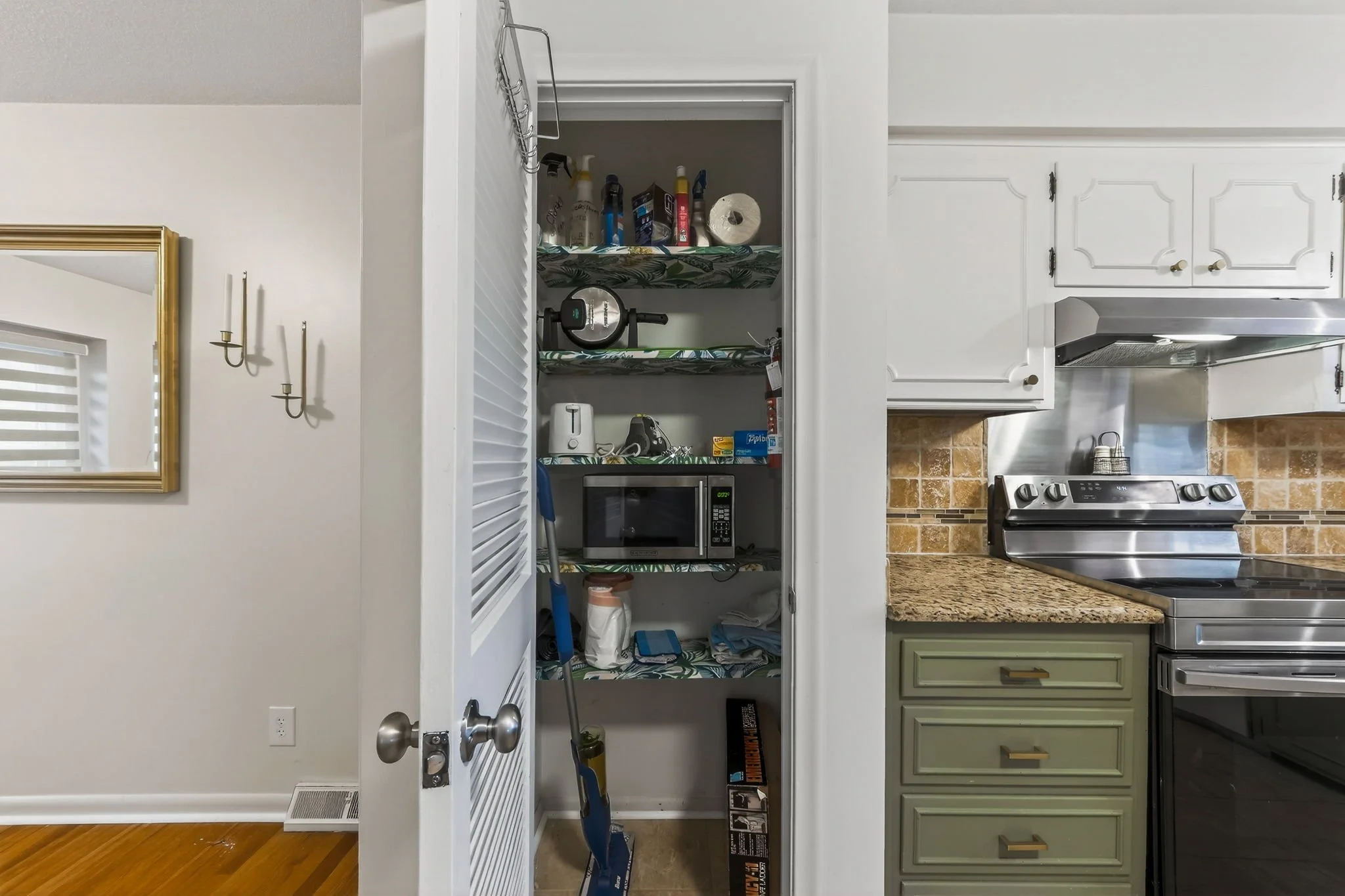 Kitchen pantry with shelves containing small appliances, cleaning supplies, and miscellaneous items, adjacent to a stove and granite countertop.