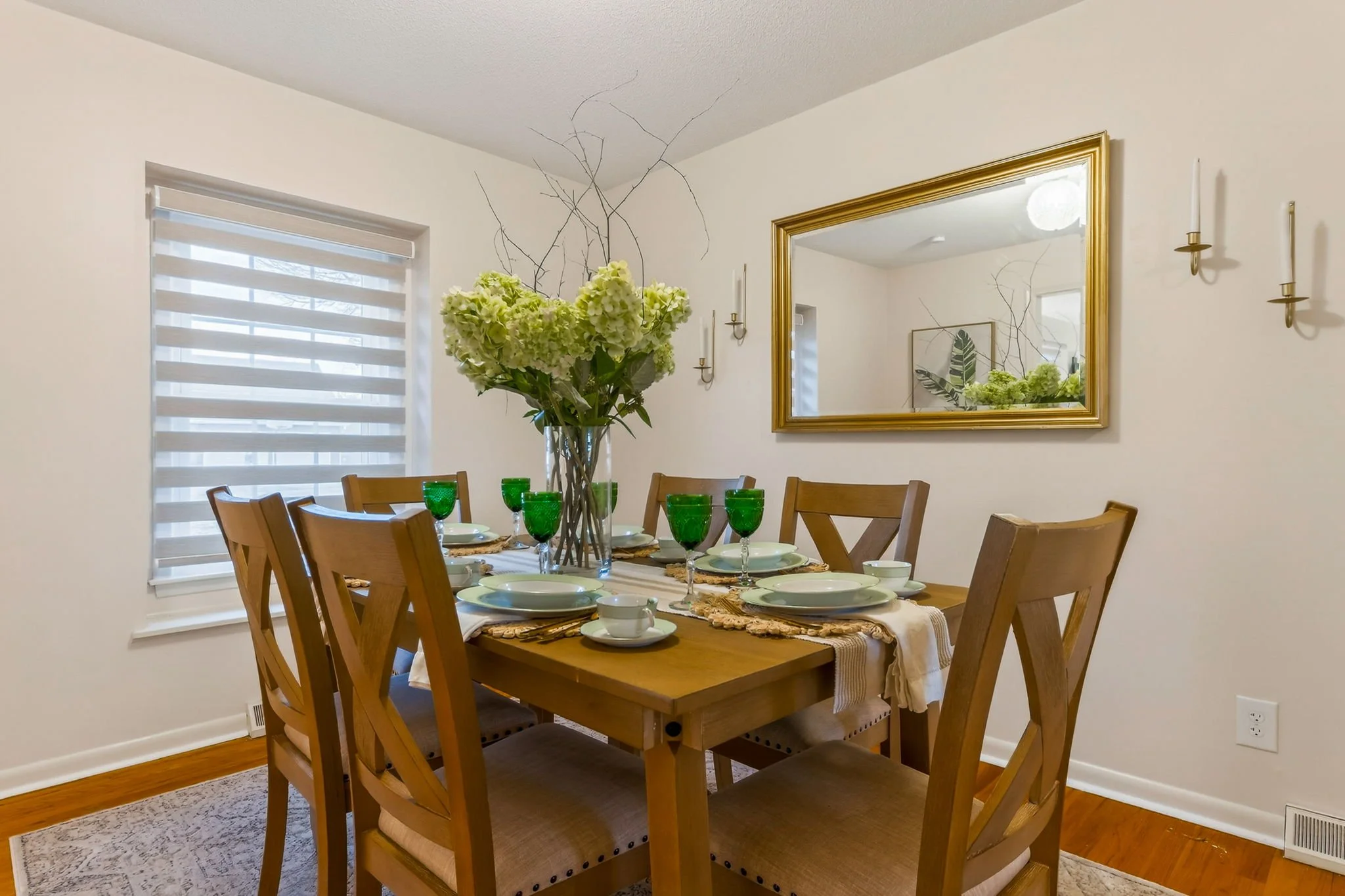 Dining room with a wooden table set for a meal, featuring white plates, green glasses, and white cups, with a large flower arrangement in the center, and a gold-framed mirror reflecting the room's decor.