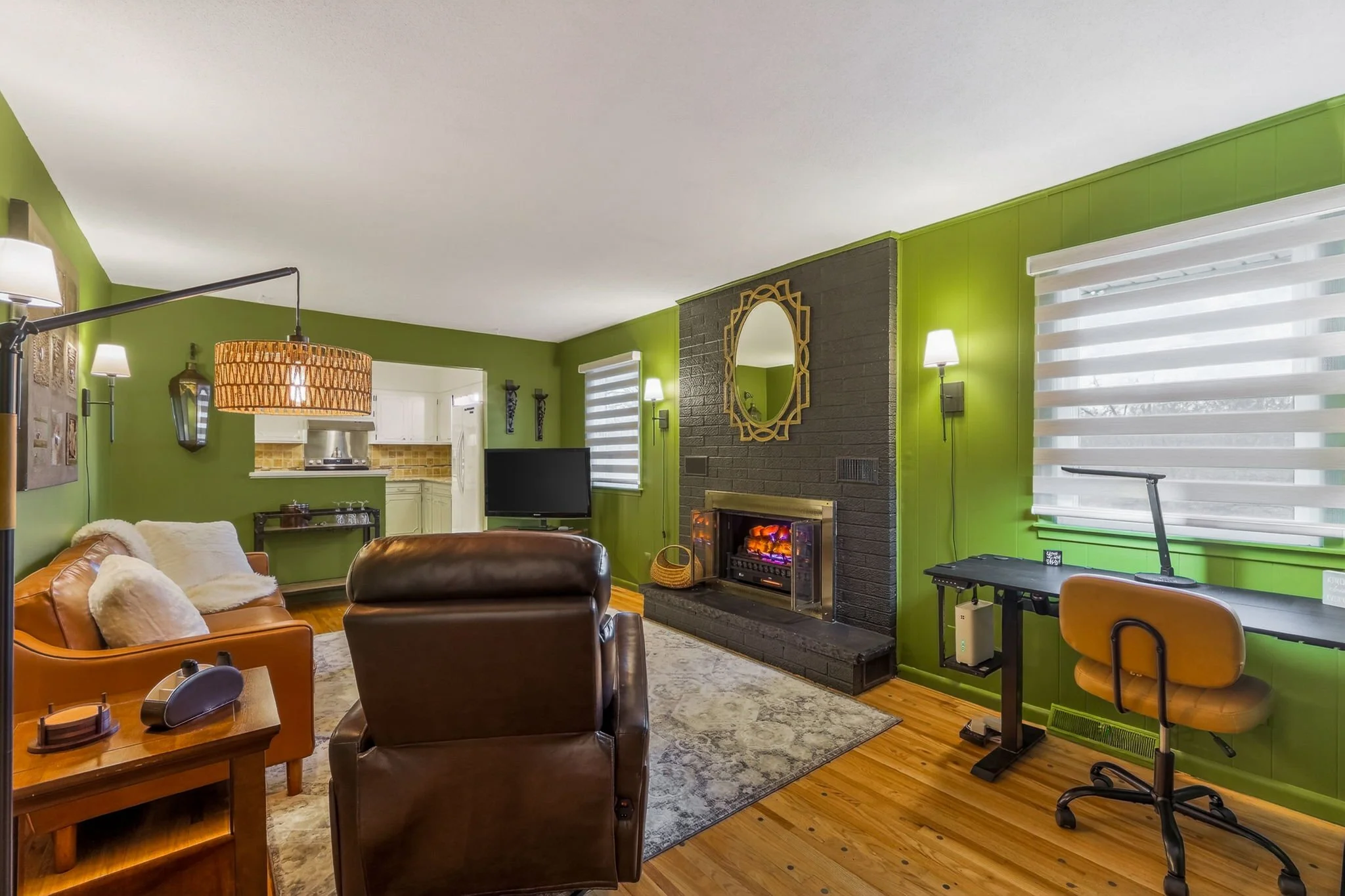 Living room with green walls, black brick fireplace, brown leather chairs, orange armchair with white pillows, TV, wooden floor, patterned rug, windows with white blinds, and a desk with a yellow chair.