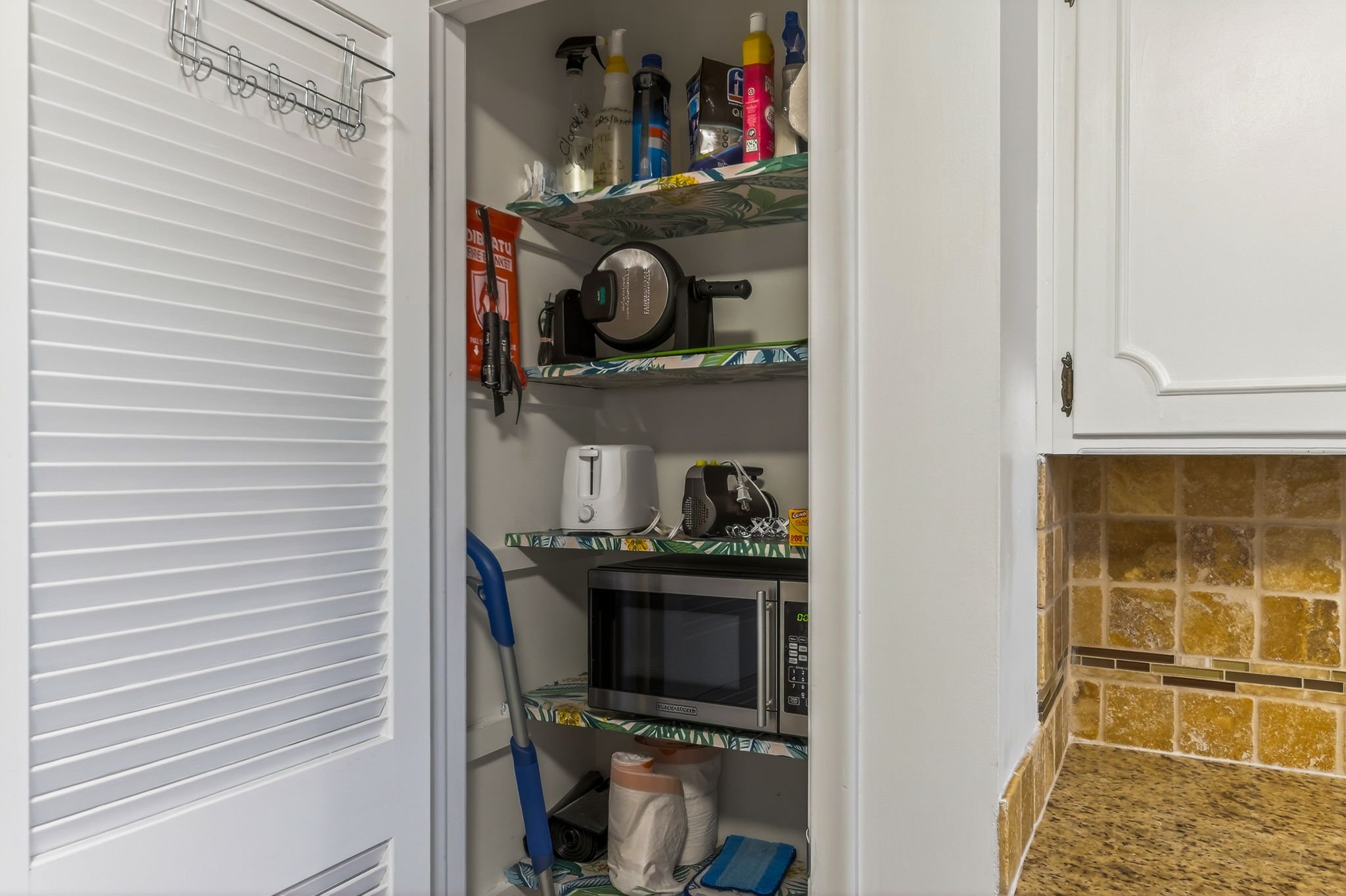 Kitchen pantry with shelves holding miscellaneous items including a microwave, toaster, kitchen tools, cleaning supplies, and a cart with a white bag and paper towels, adjacent to a kitchen counter with a tile backsplash.