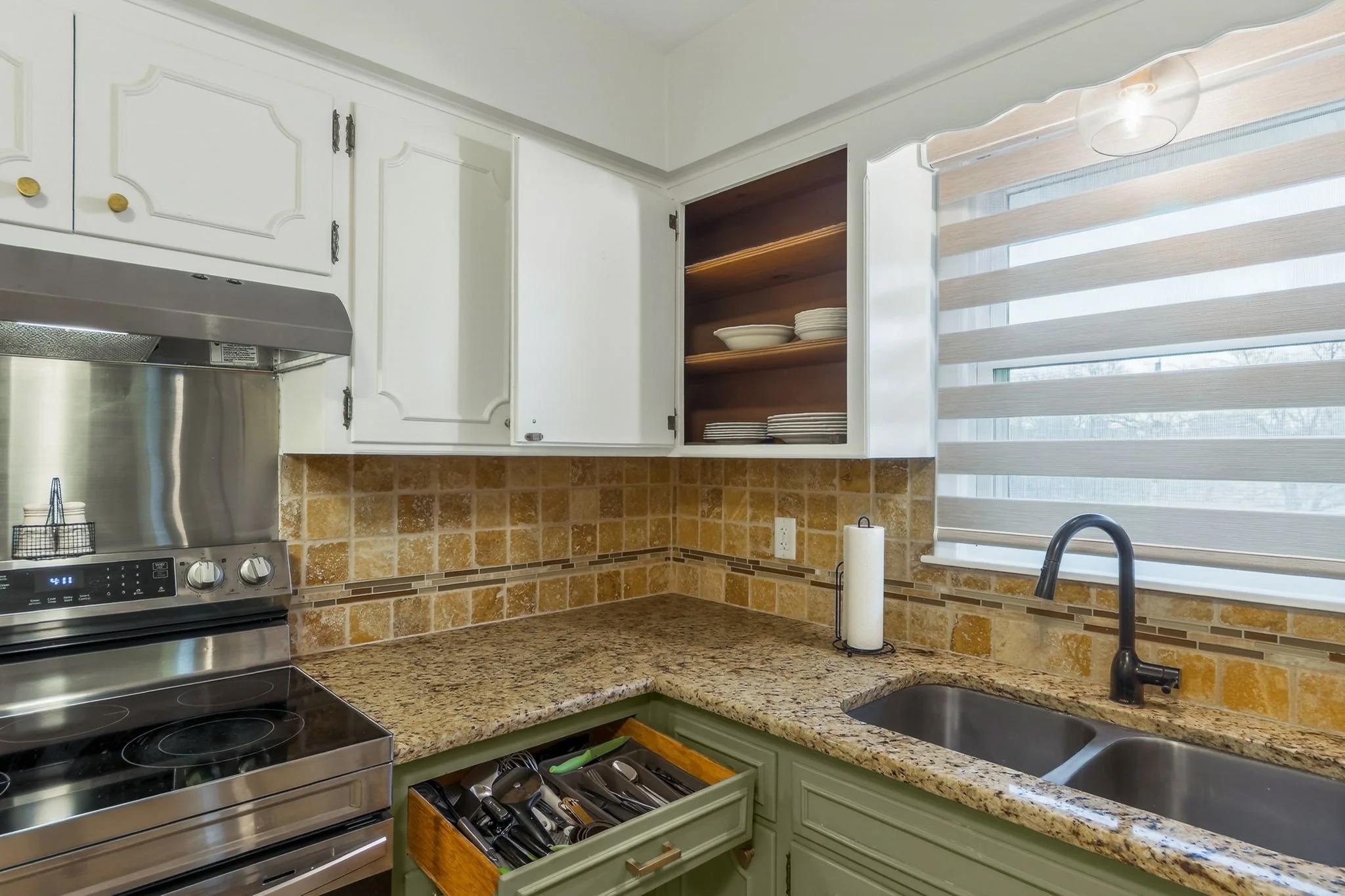 Kitchen with white cabinets, a granite countertop, a double sink, a paper towel holder, and a window with striped shades. Open cabinets show dishes inside. There is a stove and a drawer with utensils.