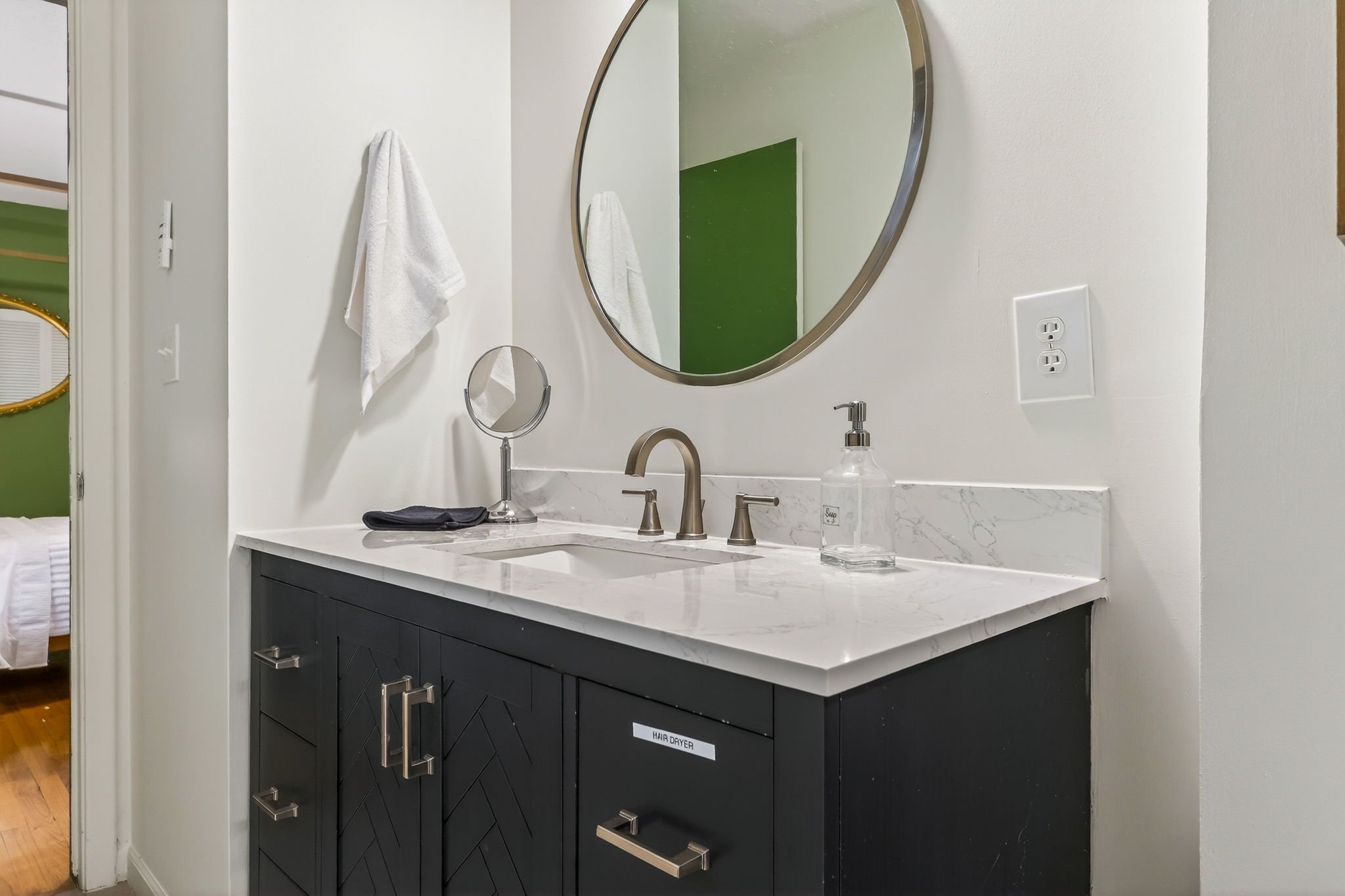 Bathroom vanity with a black cabinet, white marble countertop, a gold-finish faucet, a round mirror above, a hand soap dispenser, and a towel hanging on the wall.