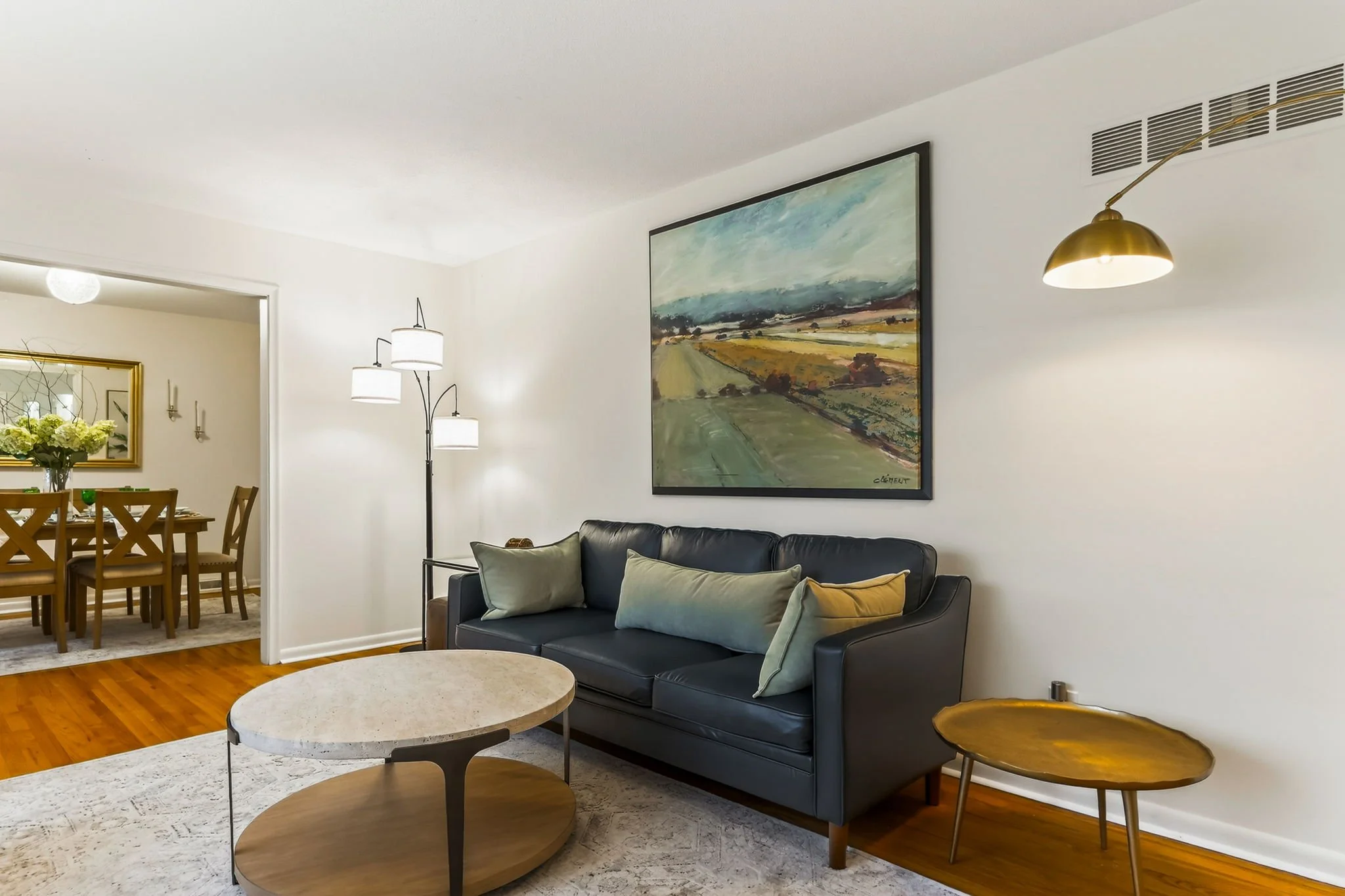 Living room with a black leather sofa, a light-colored pillows, a round marble-top coffee table, a wooden side table, a large painting of a landscape on the wall, a floor lamp with three white shades, and a gold-colored wall sconce. A dining area wit