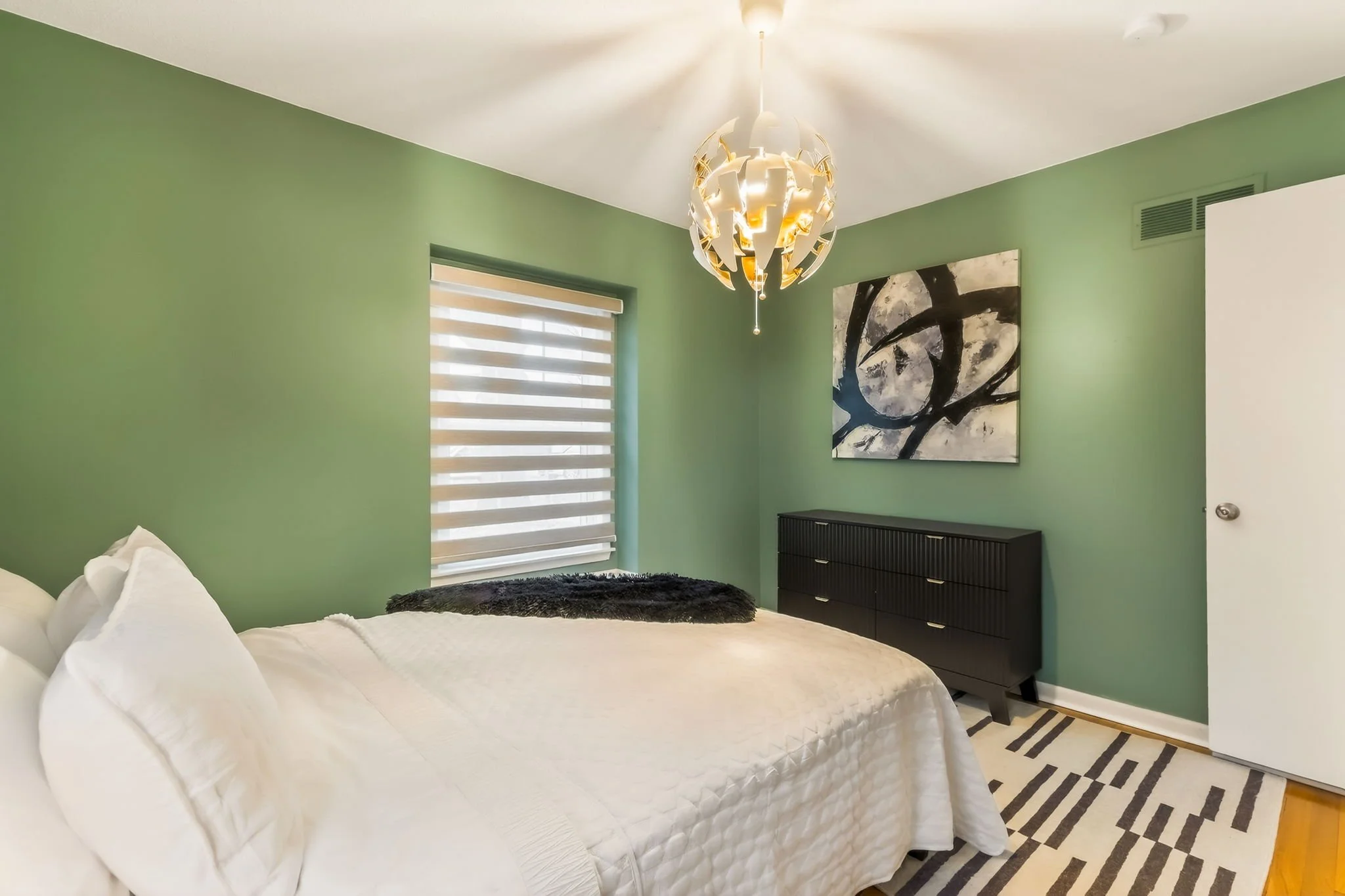 A bedroom with green walls, a white bed with pillows and a blanket, a window with striped blinds, a black dresser, an abstract black and white painting, a modern white chandelier, and a patterned rug.