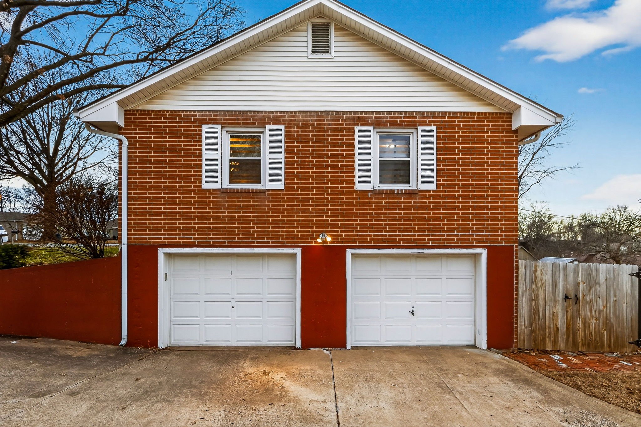 A two-story house with a brick facade, white shutters on the windows, two white garage doors, and a white gable roof, with a concrete driveway and a wooden fence on the right side.