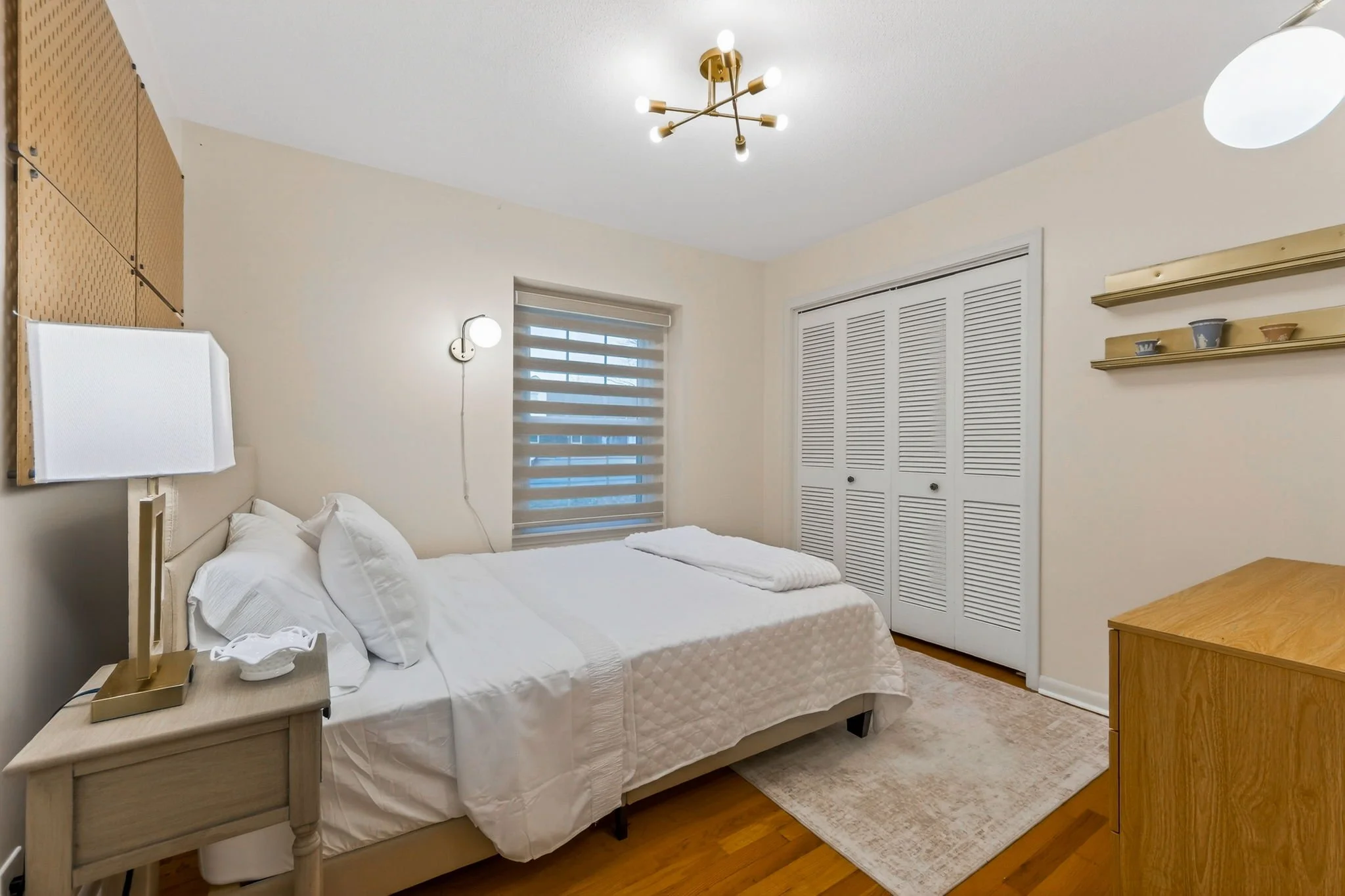 A bedroom with a bed covered in white bedding and pillows, a nightstand with a lamp, a window with blinds, a closet with louvered doors, and wooden shelves with small decorative items.