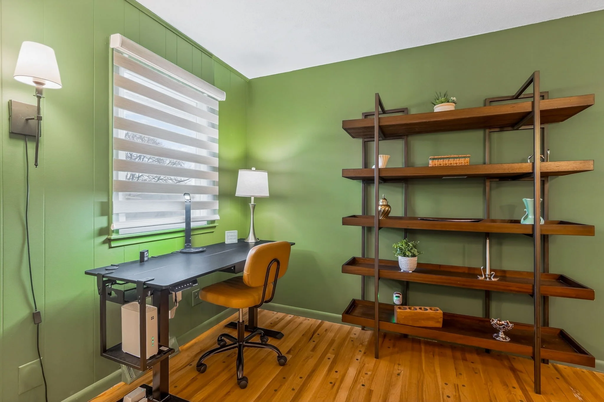 Home office with green walls, a black desk, a yellow office chair, a window with a white blind, a standing lamp, a bookshelf with plants and decorative items, and hardwood floors.