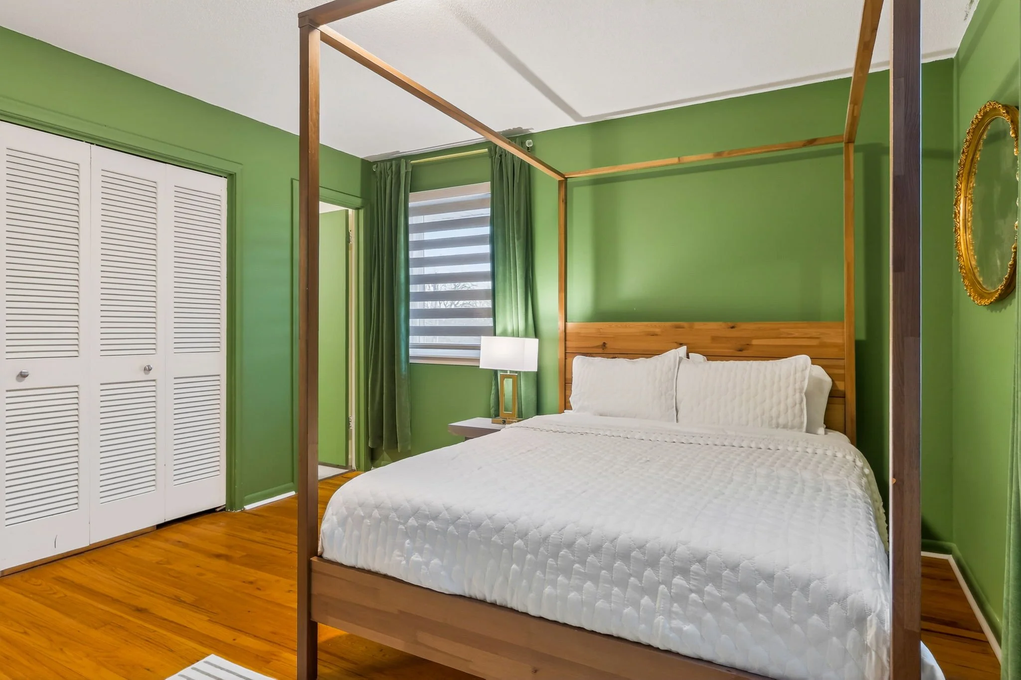 Bedroom with green walls, a wooden four-poster bed with white bedding, a white nightstand with a lamp, a window with striped blinds and green curtains, wooden flooring, and a gold-framed oval mirror on the wall.