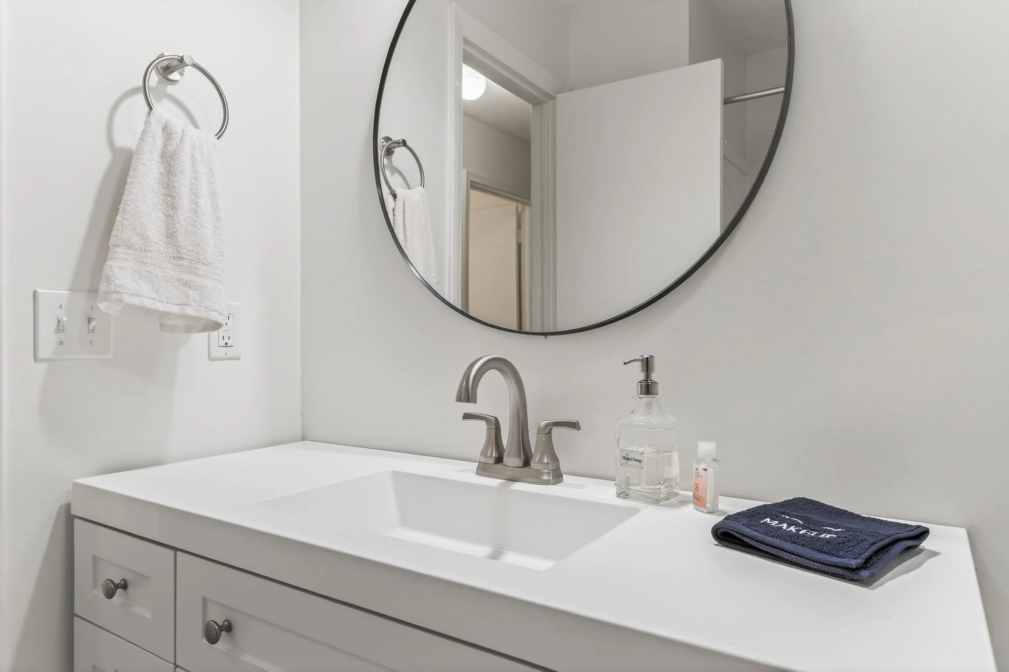 Bathroom sink with a chrome faucet, a round mirror, and a white vanity with two drawers. There is a soap dispenser, a small bottle, a folded dark blue towel, a white hand towel on a towel ring, and a wall socket.