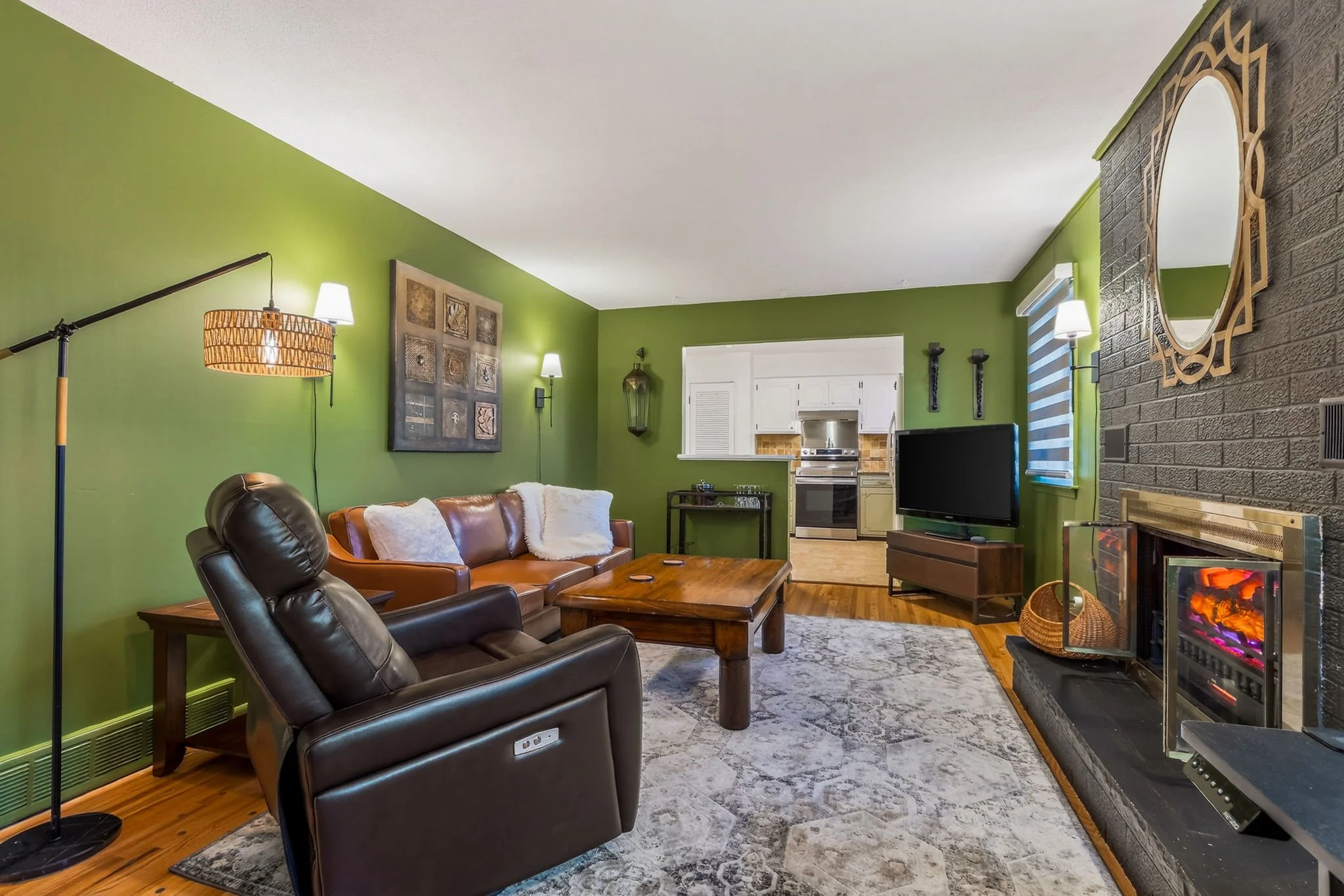 Living room with green walls, leather recliner, sofa with pillows, wooden coffee table, TV on a stand, fireplace with lit fire, and artwork and mirrors on the walls. Open kitchen in the background.