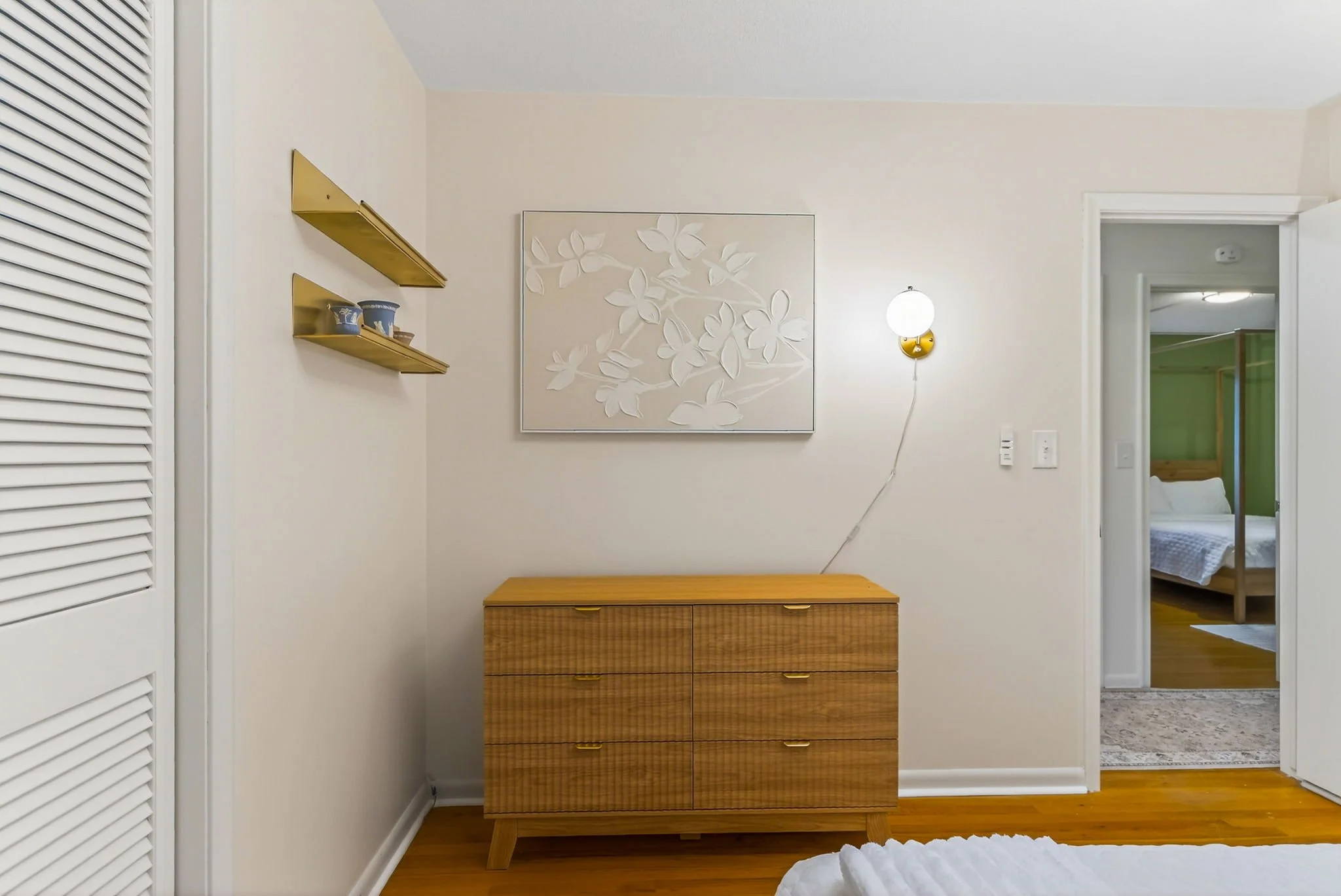 A bedroom featuring a wooden dresser, three gold shelves with small blue patterned containers, a wall sconce with a white globe, a framed white floral art piece, white walls, and a door leading to a second bedroom with a green accent wall and a woode