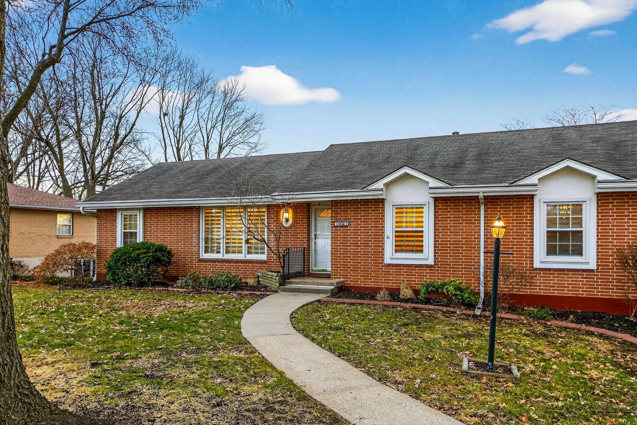 Front view of a brick single-story house with white trim, three windows with shutters, a front door, and a curved concrete walkway leading to the entrance. There are leafless trees in the background and a street lamp lighting the lawn.